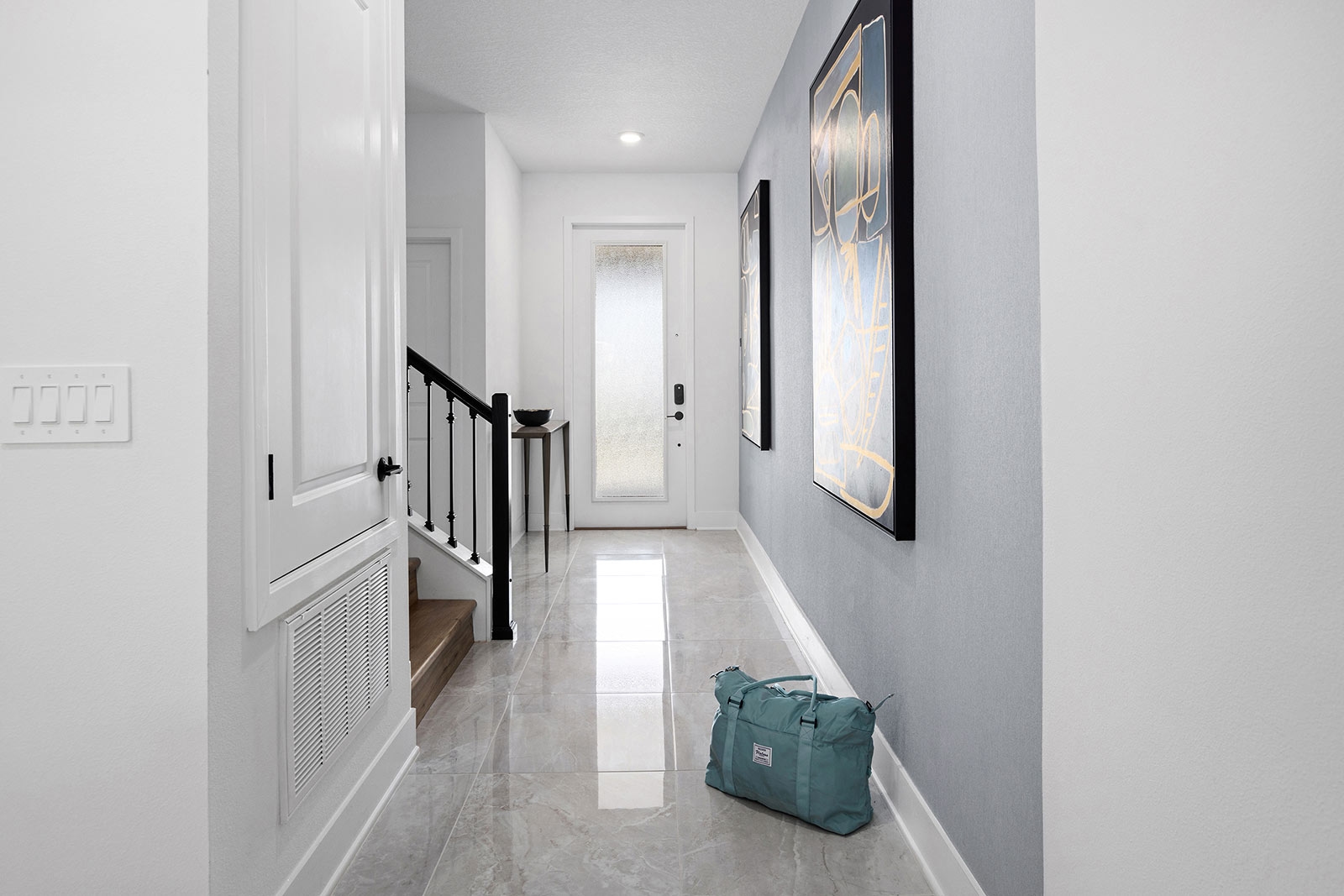 A bright villa entryway featuring polished marble-style tile floors, a light blue accent wall decorated with abstract art, a wooden staircase, and a glass-paneled front door, with a blue duffel bag sitting on the floor.