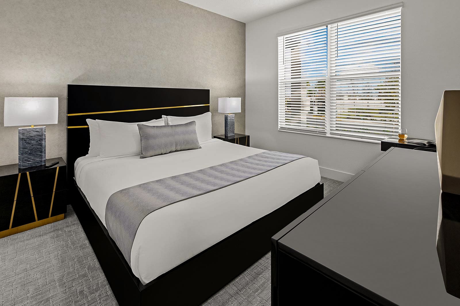 A modern bedroom featuring a king bed with a black and gold headboard, a textured accent wall, and a large window with blinds.