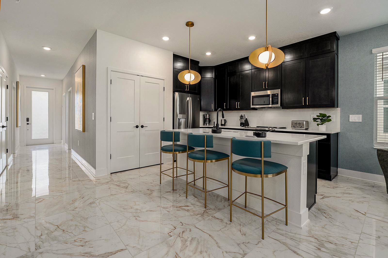 A contemporary kitchen with dark cabinetry, white marble-style tile floors, and a white island counter paired with three teal and gold barstools.