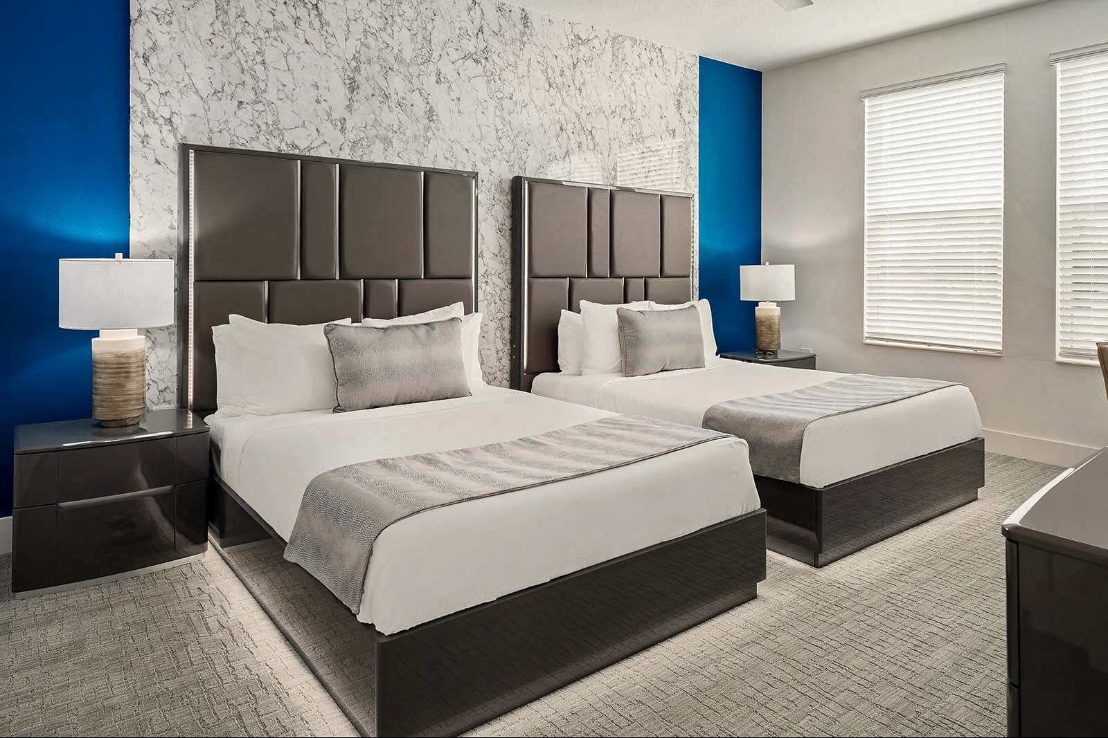 A modern bedroom featuring two queen beds with dark brown tufted headboards against a white marble-patterned accent wall flanked by vibrant blue panels.