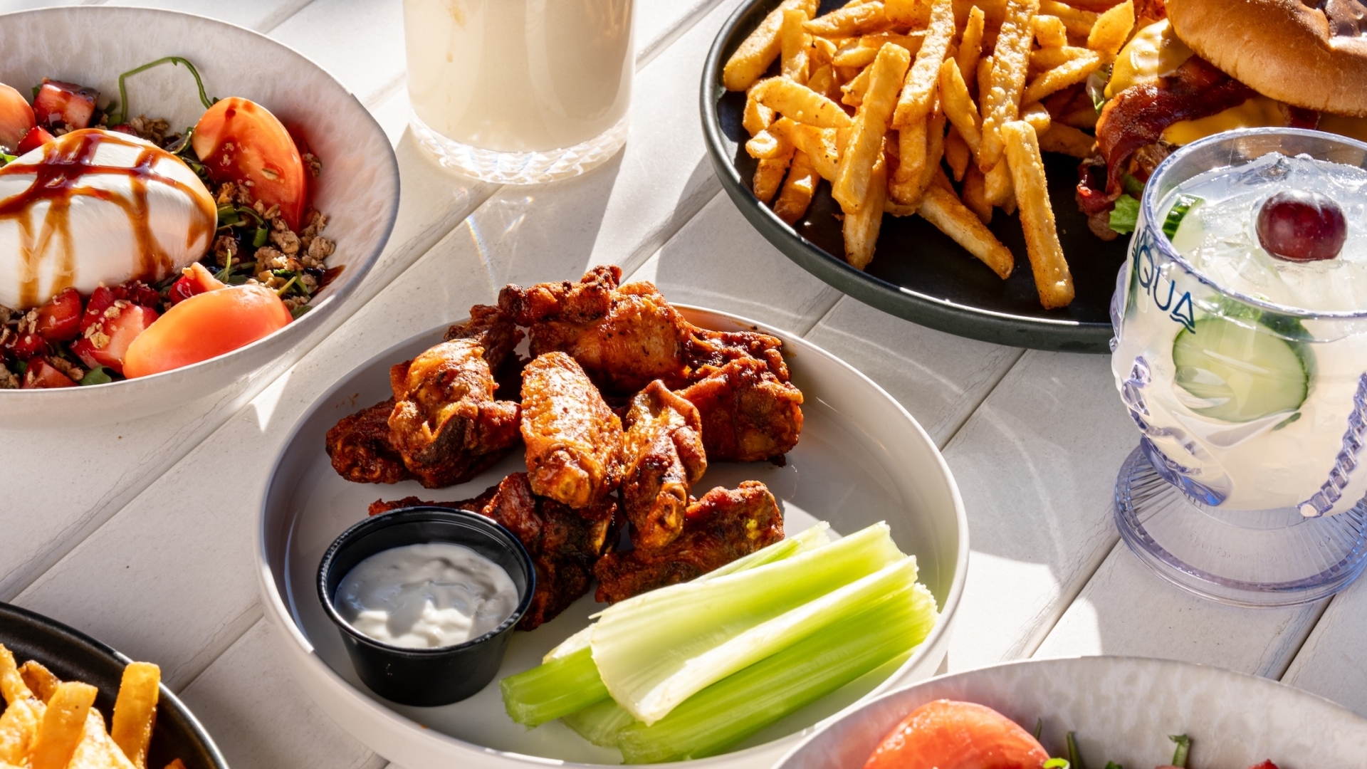 A variety of appetizers and entrees, including buffalo wings and a fresh strawberry burrata salad, arranged on a white outdoor table.