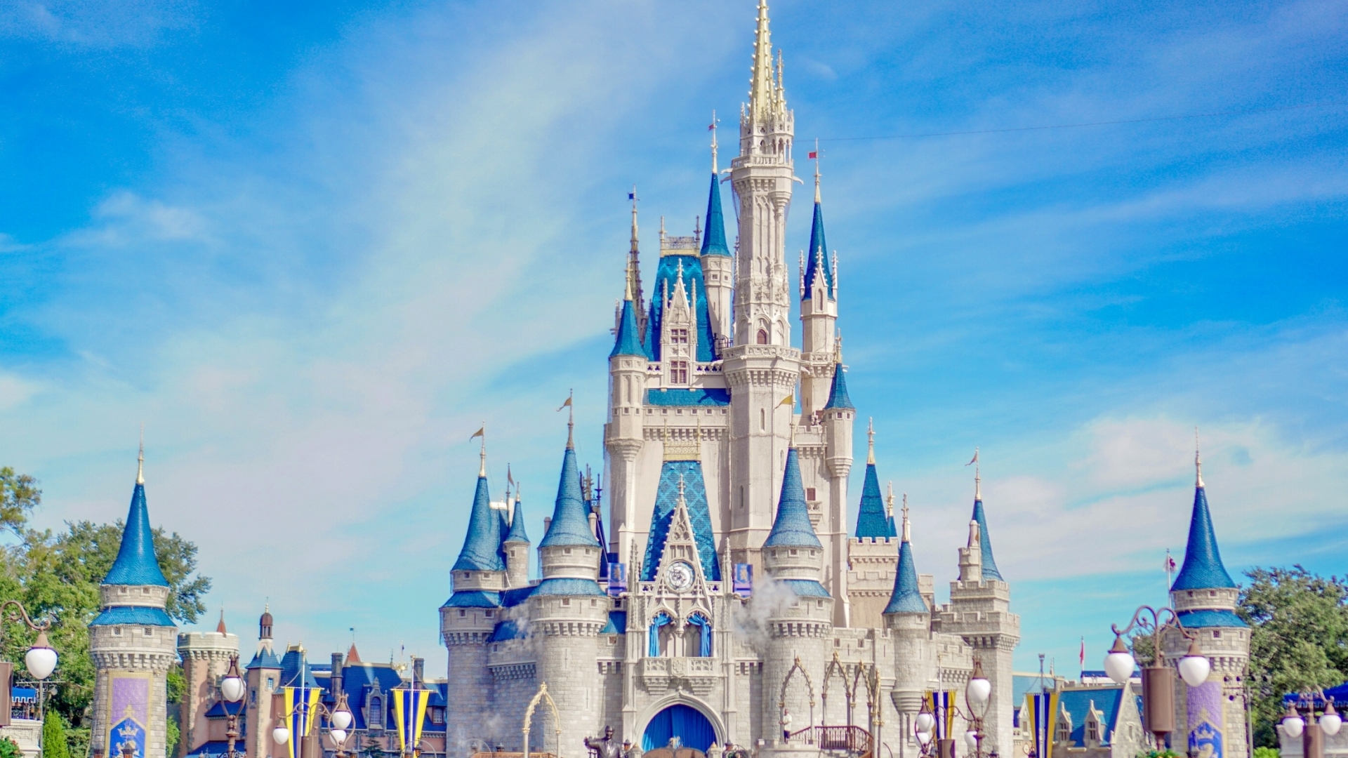 A wide view of Cinderella Castle at Magic Kingdom Park under a bright blue sky, featuring iconic blue and gold spires and the "Partners" statue of Walt Disney and Mickey Mouse in the foreground.
