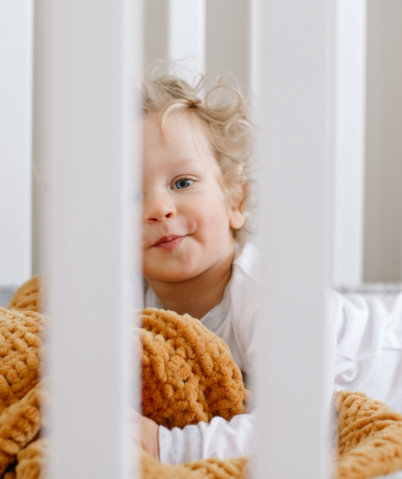 Close-up of a smiling toddler peeking through the white slats of a rented wooden crib with a soft yellow blanket.