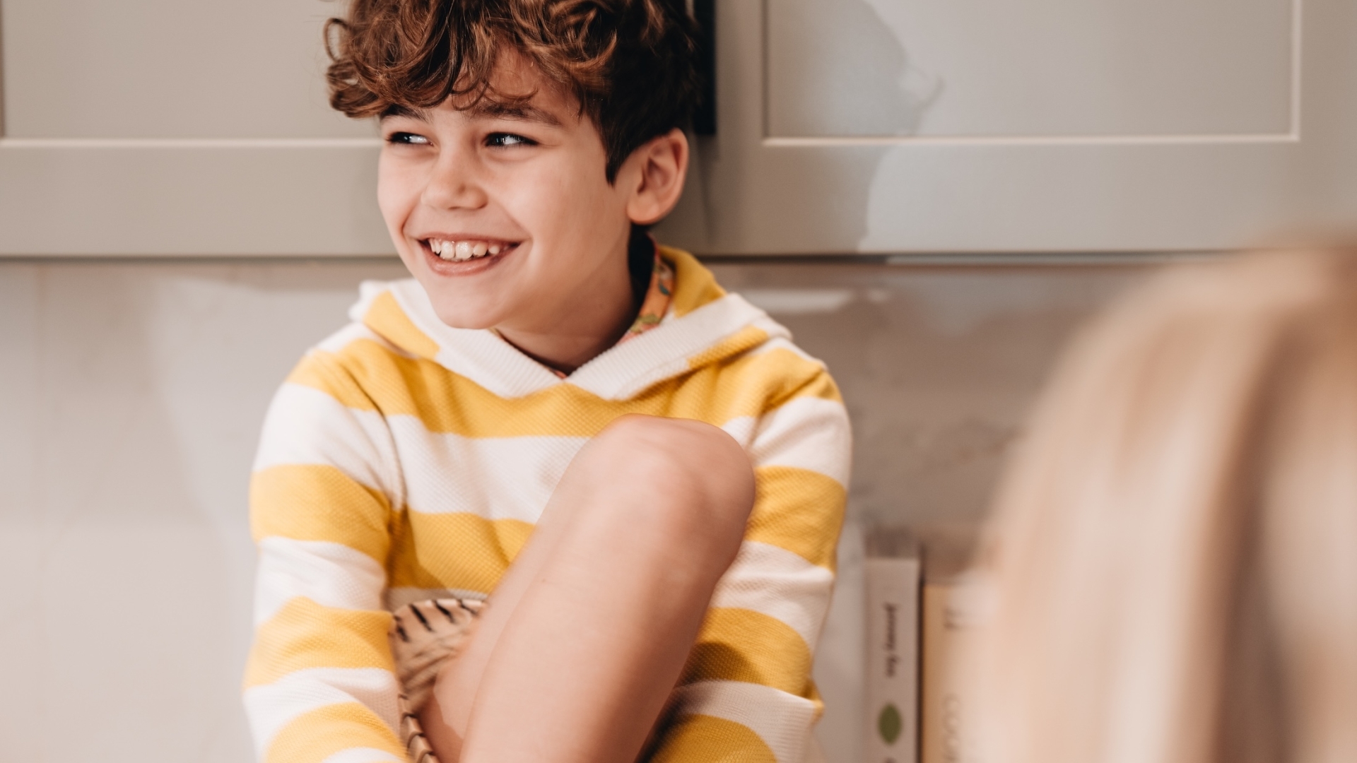A candid close-up of a young boy with curly hair laughing and wearing a yellow and white striped hoodie.