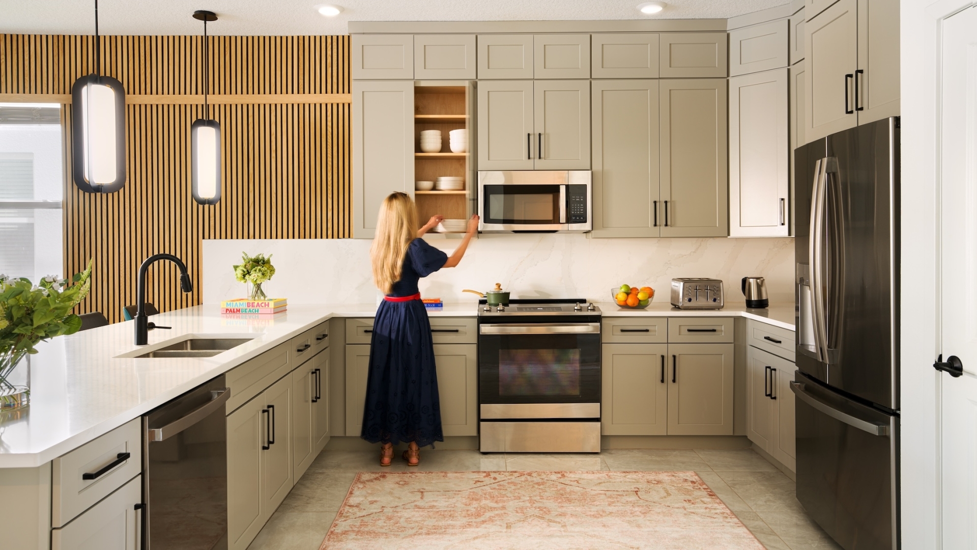 A woman in a blue dress organizing dishes inside a spacious contemporary kitchen featuring double cabinetry, quartz countertops, and a large pink patterned rug.