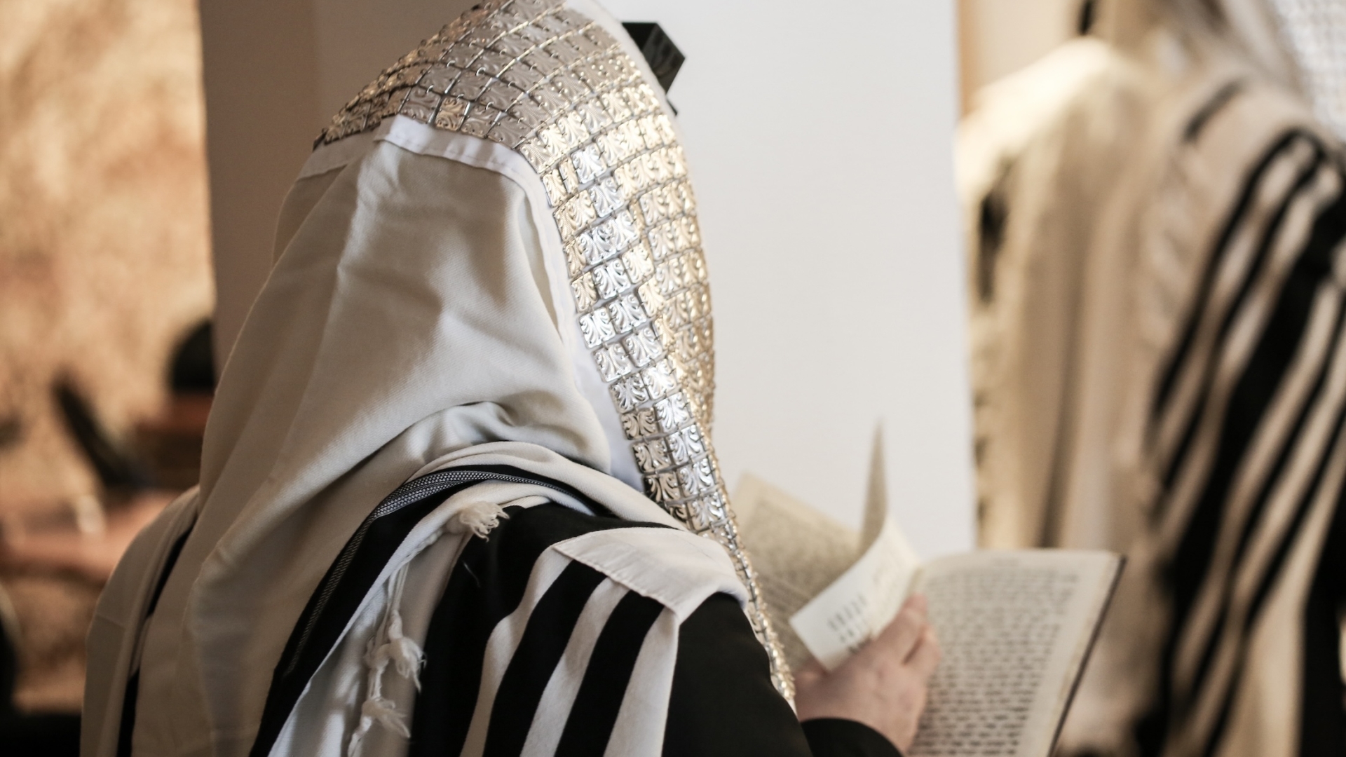 A close-up, over-the-shoulder view of a Jewish man wearing a traditional tallit prayer shawl with an ornate silver atarah band, reading from a prayer book during a service.
