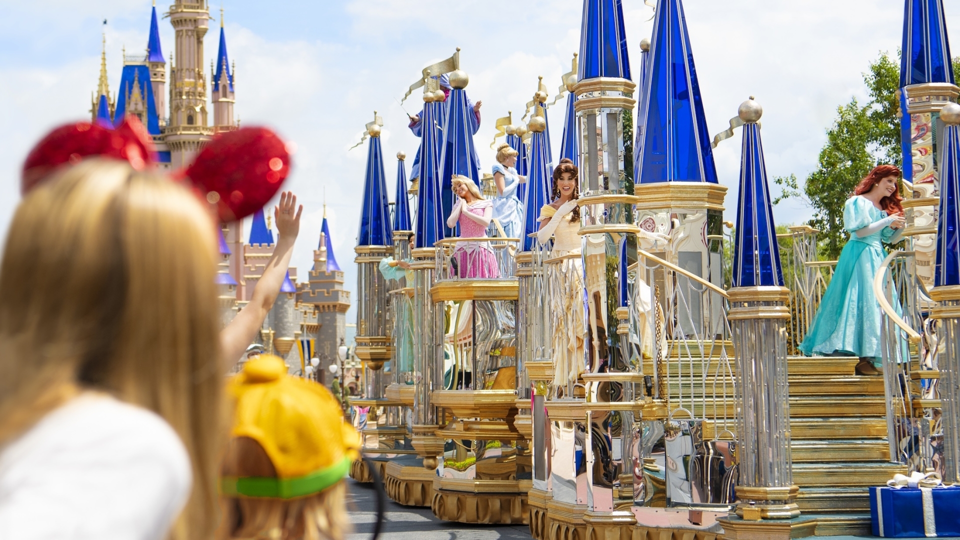 Disney characters on a blue and gold parade float in front of Cinderella Castle at Magic Kingdom, with a child in the foreground waving.