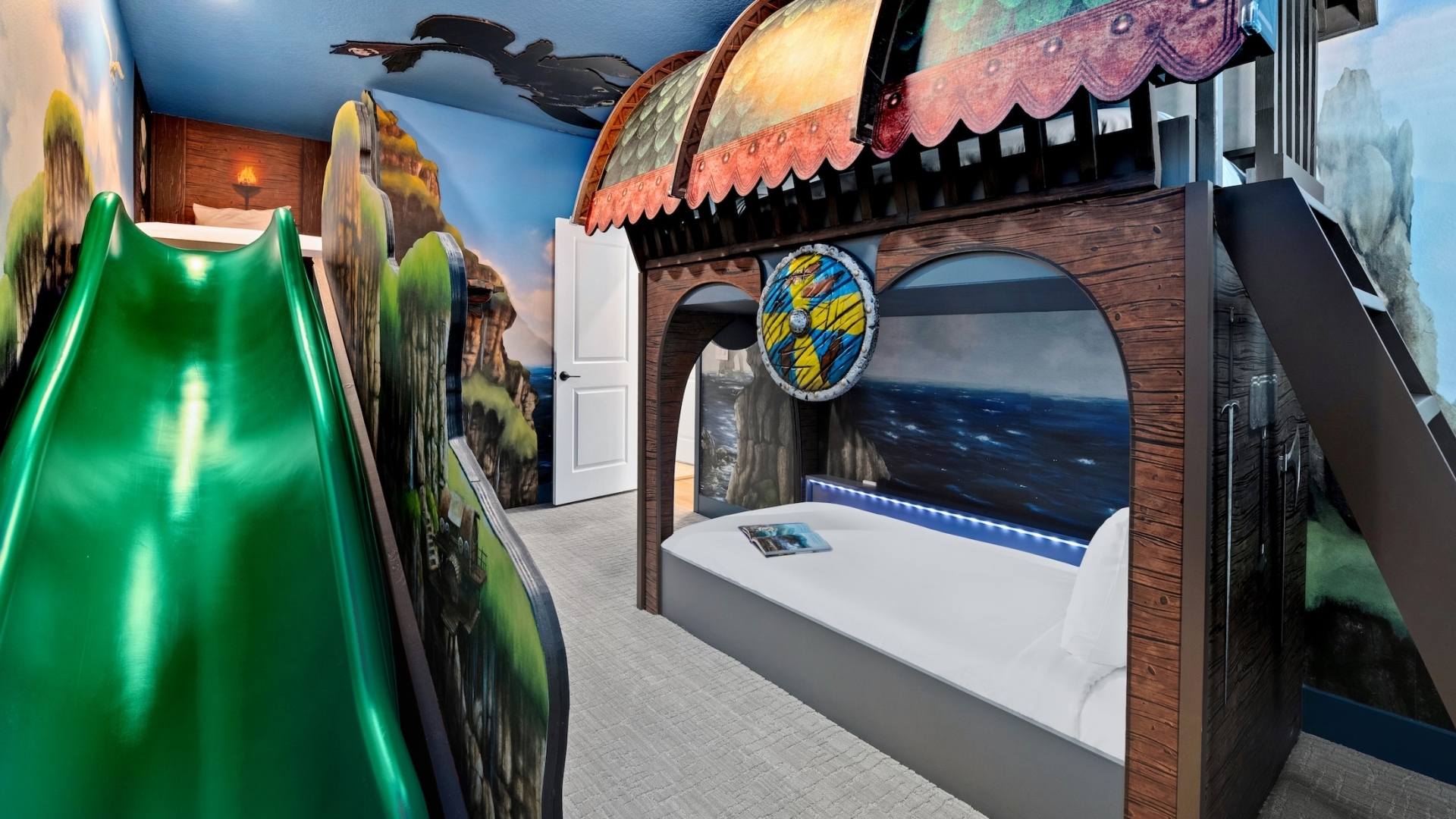 A fantasy-themed children's bedroom featuring a Viking ship bunk bed with a decorative shield, a green slide, and immersive murals of dragons and coastal mountains.