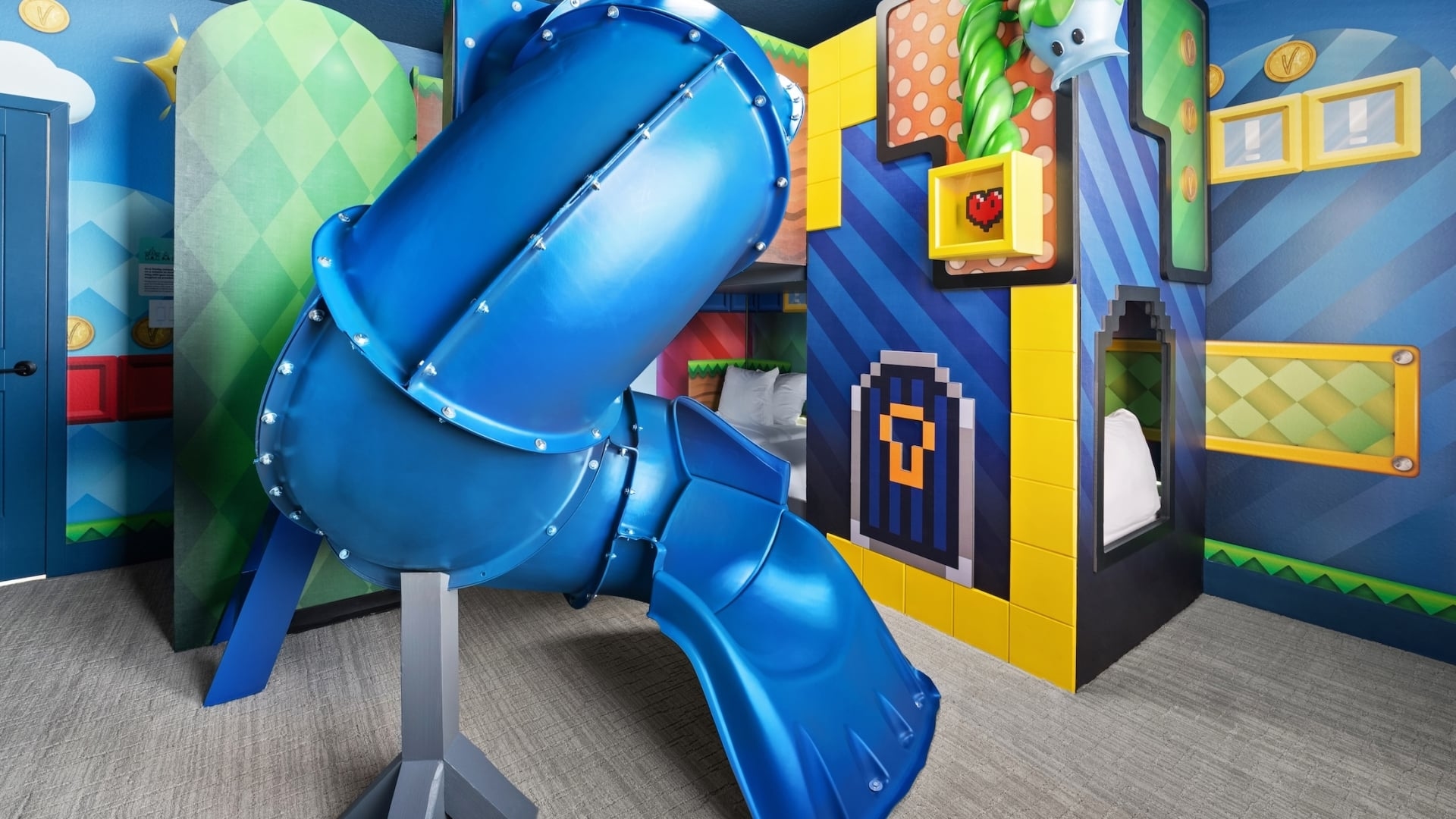 A video-game-themed children's bedroom featuring pixel-art wall murals, a bunk bed designed as a game console, and a large blue winding tunnel slide.