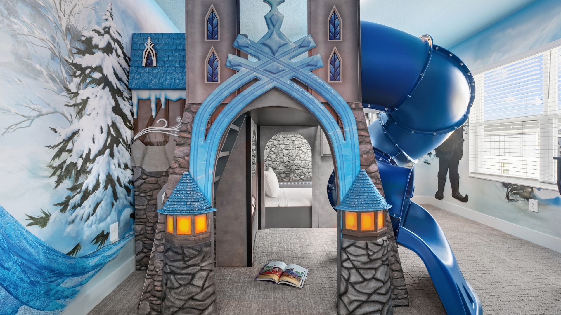 An ice-castle-themed children's bedroom featuring a gray stone bunk bed structure with blue turrets, a winding blue tunnel slide, and snowy forest wall murals.