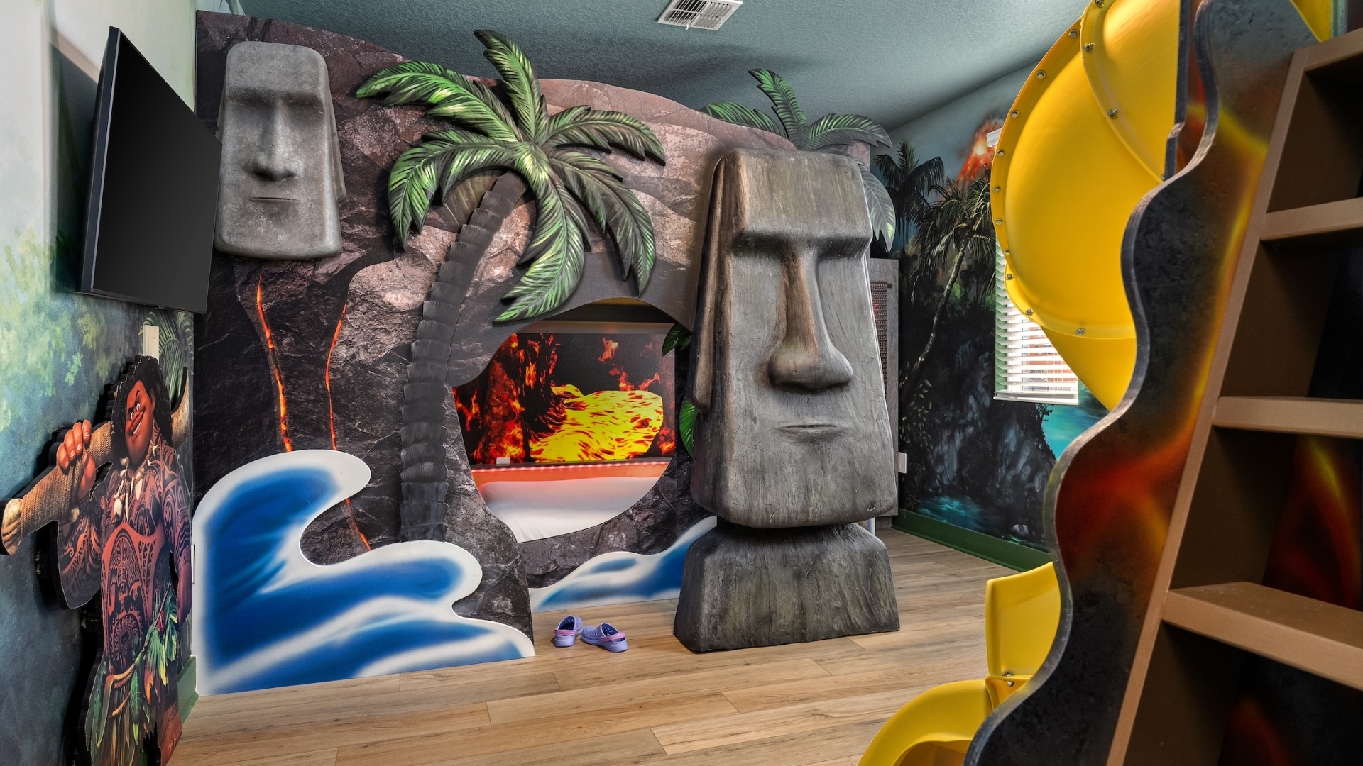 A tropical island-themed children's bedroom featuring Moai statue structures, a bunk bed with a lava mural, a winding yellow slide, and palm tree decor.