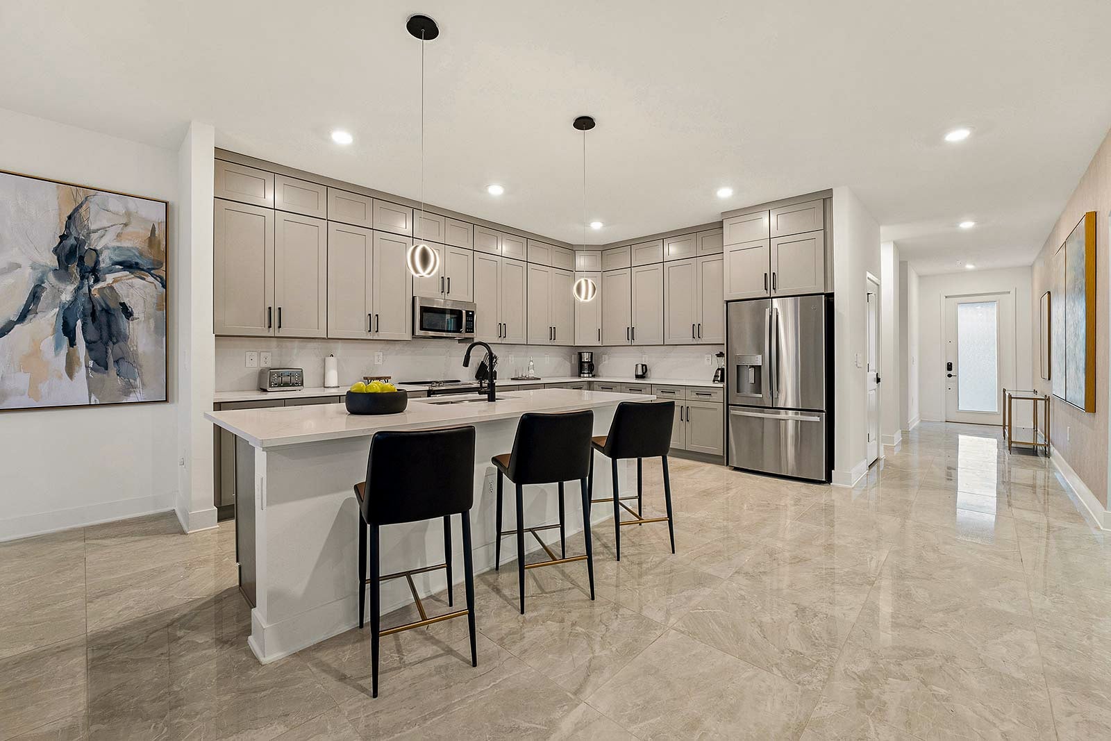A spacious kitchen featuring minimalist light gray cabinets, a white quartz island with three black leather barstools, and polished tile flooring.