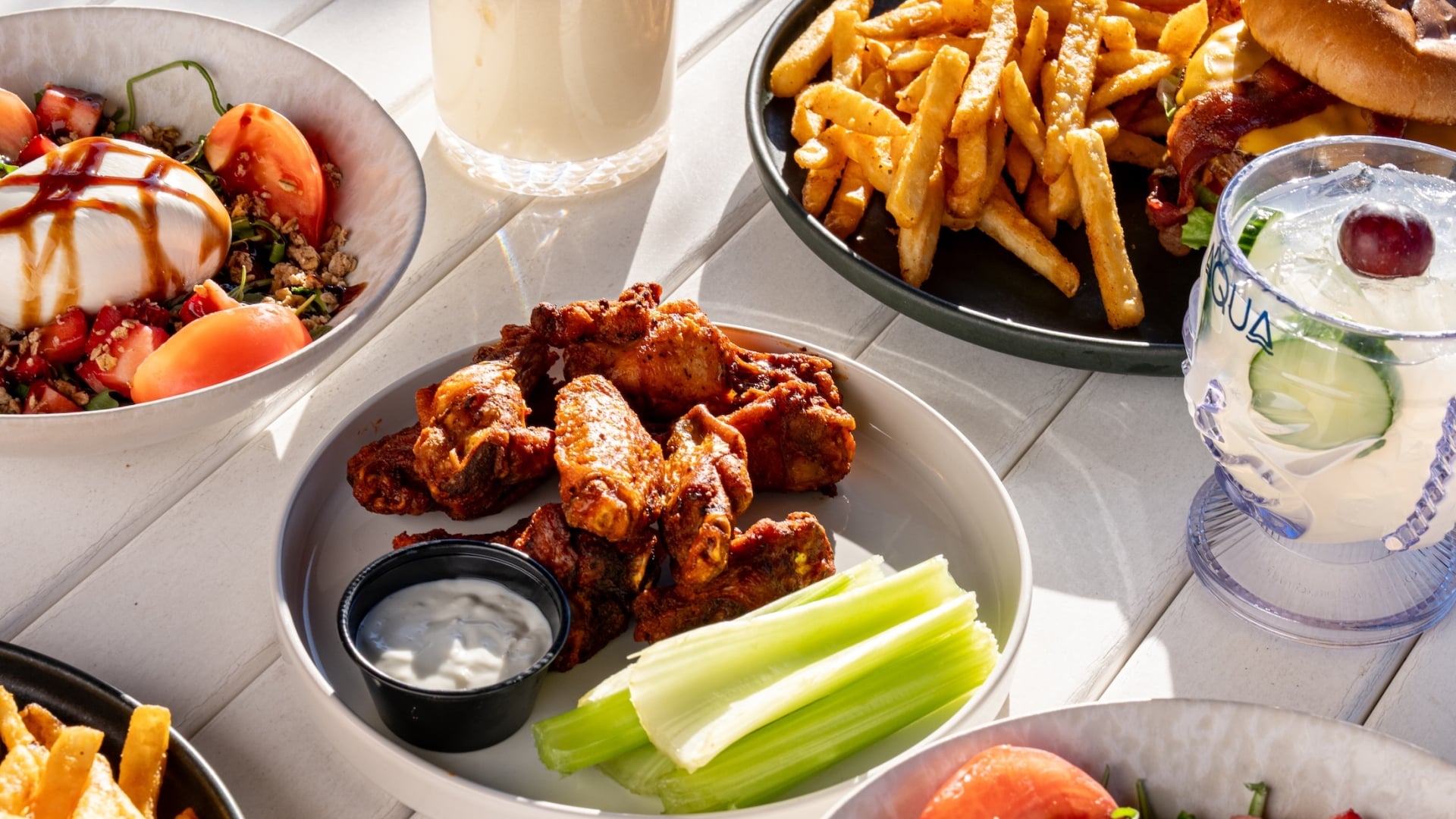 A variety of appetizers and entrees, including buffalo wings and a fresh strawberry burrata salad, arranged on a white outdoor table.