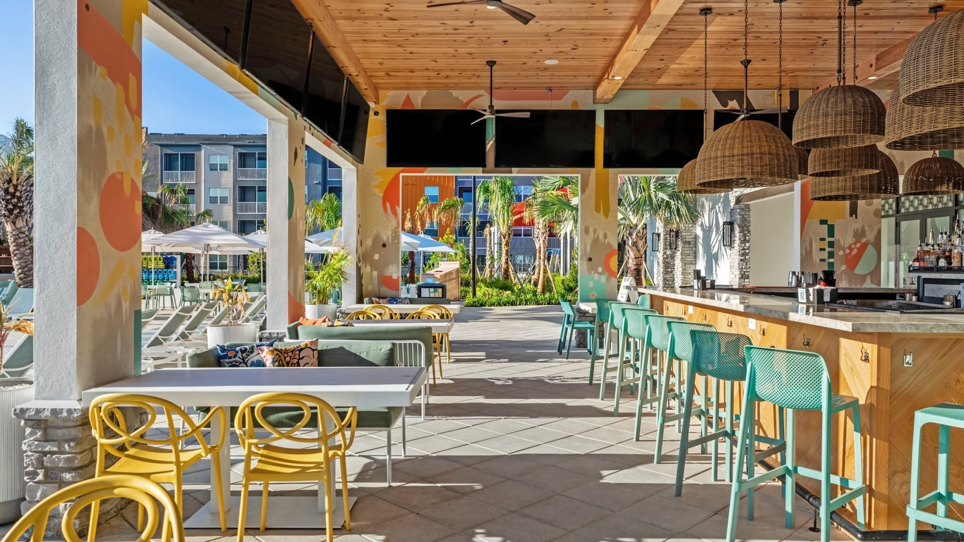 Upscale patio dining area with yellow and teal seating, wall-mounted televisions, and an open view of the resort pool deck.