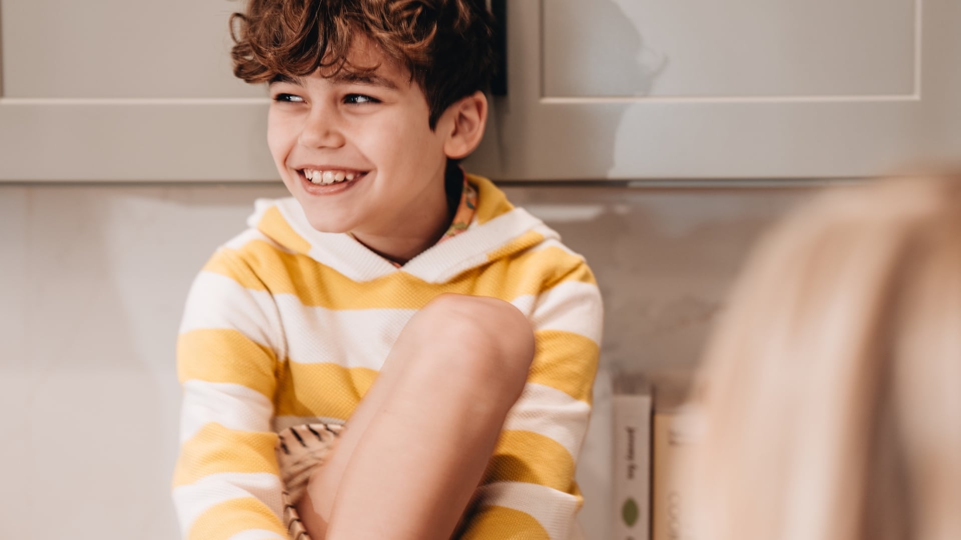 A candid close-up of a young boy with curly hair laughing and wearing a yellow and white striped hoodie.