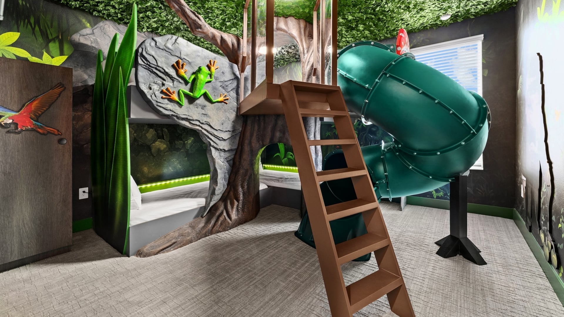 A tropical jungle-themed children's bedroom featuring a treehouse bunk bed structure with a green tunnel slide, a faux-foliage ceiling, and a large 3D frog sculpture on a rock-textured wall.