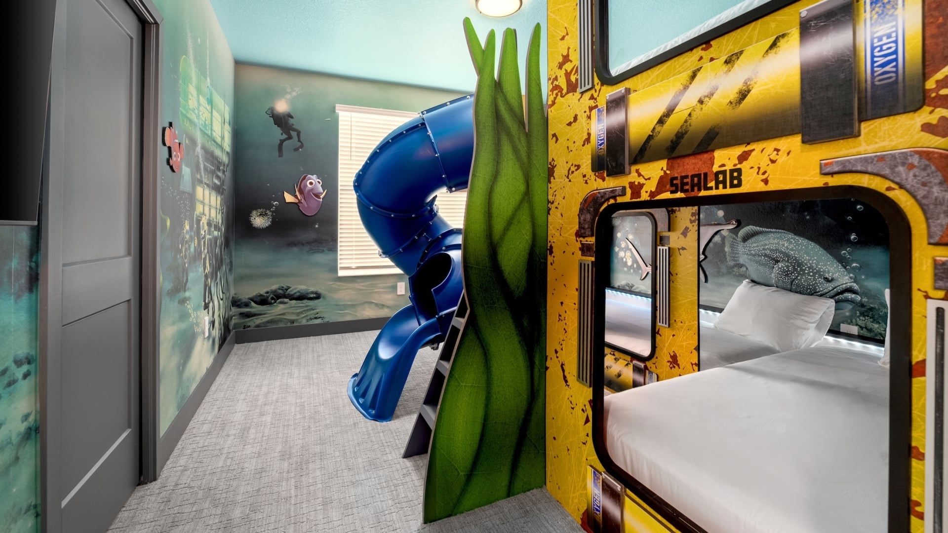 An underwater-themed children's bedroom featuring a "Sealab" industrial-style bunk bed, a blue winding tunnel slide, and immersive wall murals of a coral reef with divers and sea life.