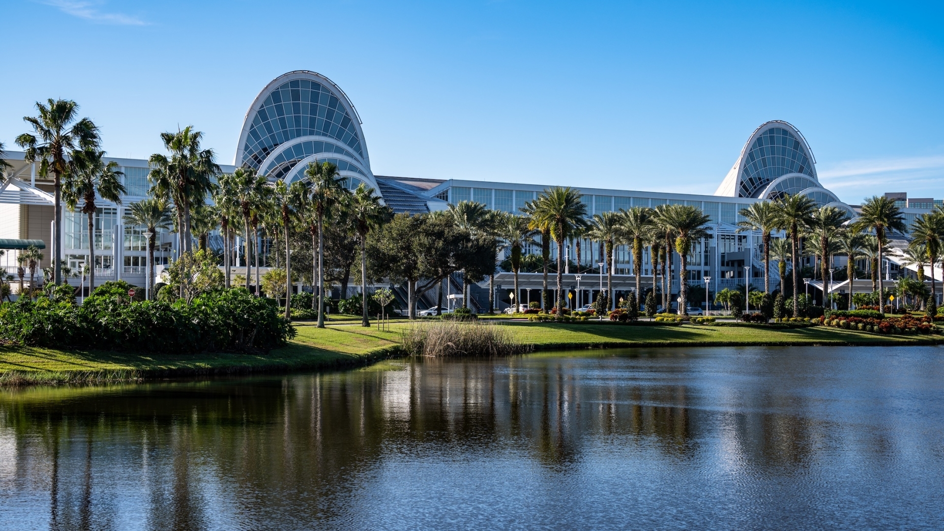 The exterior of the Orange County Convention Center in Orlando, showcasing its signature arched glass entryways and palm tree-lined waterfront landscape reflected in a calm lake under a clear blue sky.