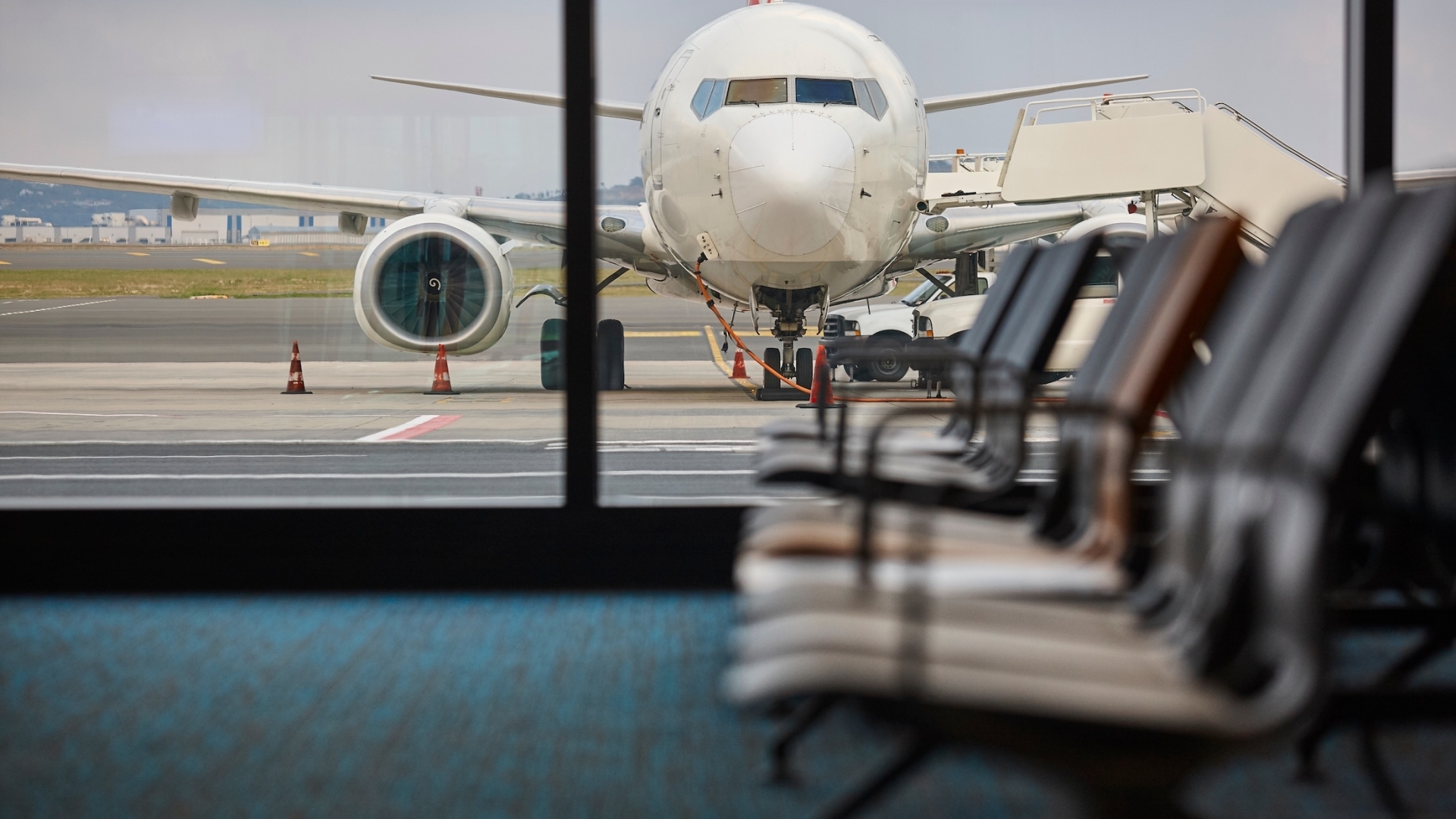 An airplane parked on the tarmac outside an airport terminal window, viewed from an indoor gate seating area with rows of chairs in the blurred foreground.