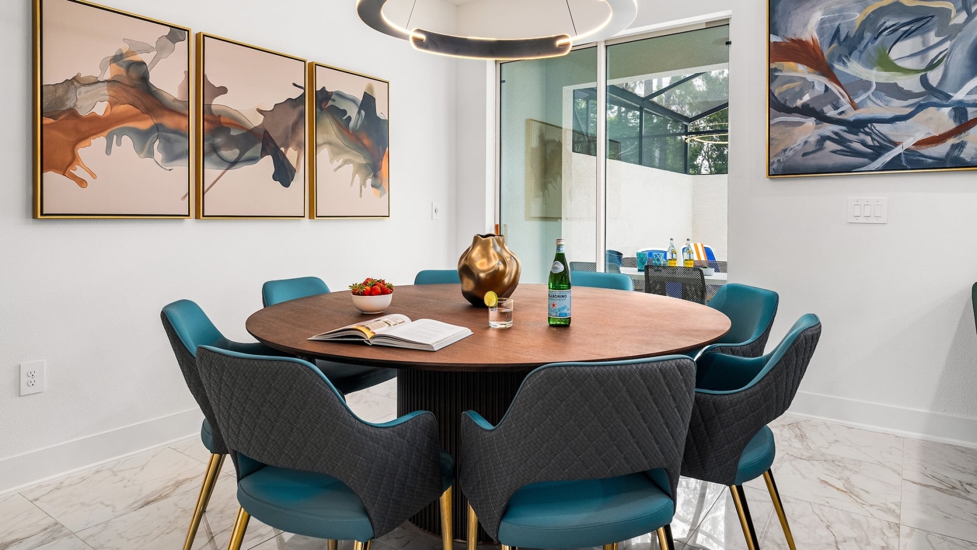 A stylish dining room with a round wooden table, teal cushioned chairs, a circular pendant light, and abstract art pieces on white walls.