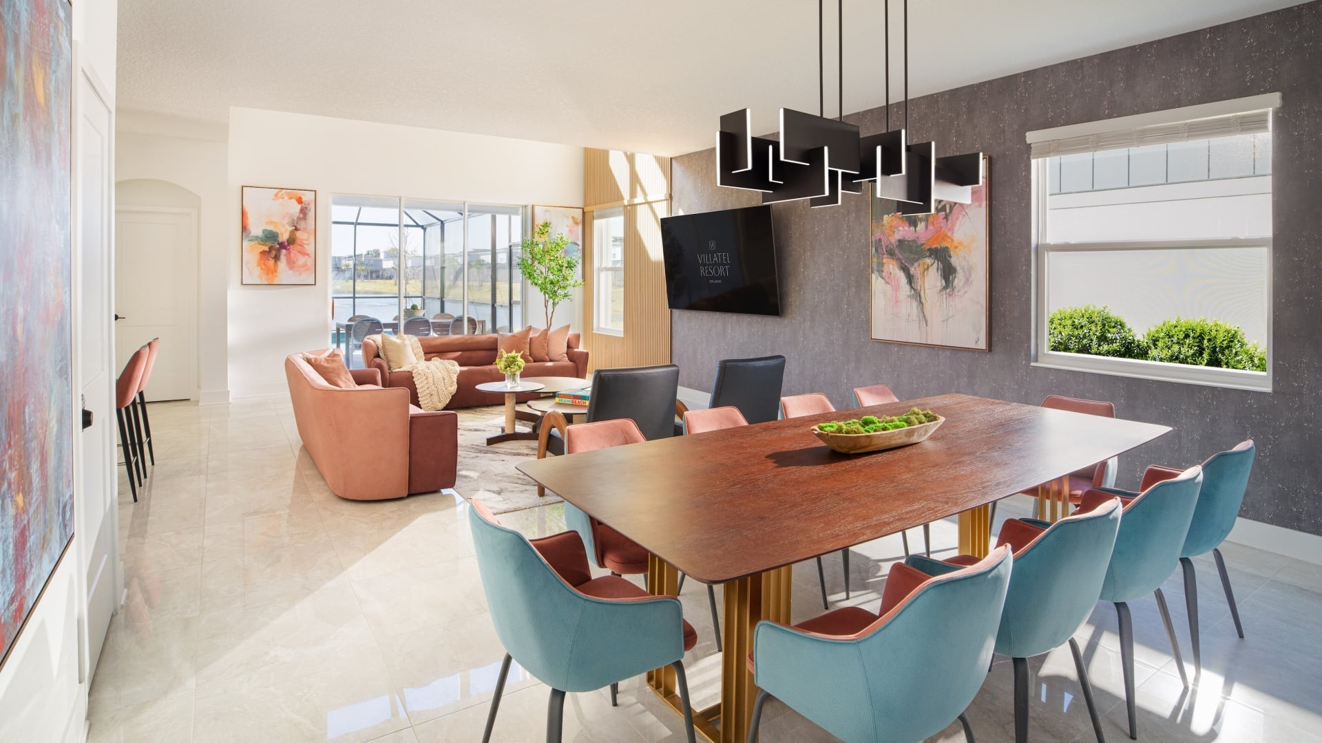 An open-concept resort villa living space featuring a large wooden dining table with dual-toned teal and pink chairs, a contemporary dark light fixture, and a living room set in the background with terracotta-colored sofas.