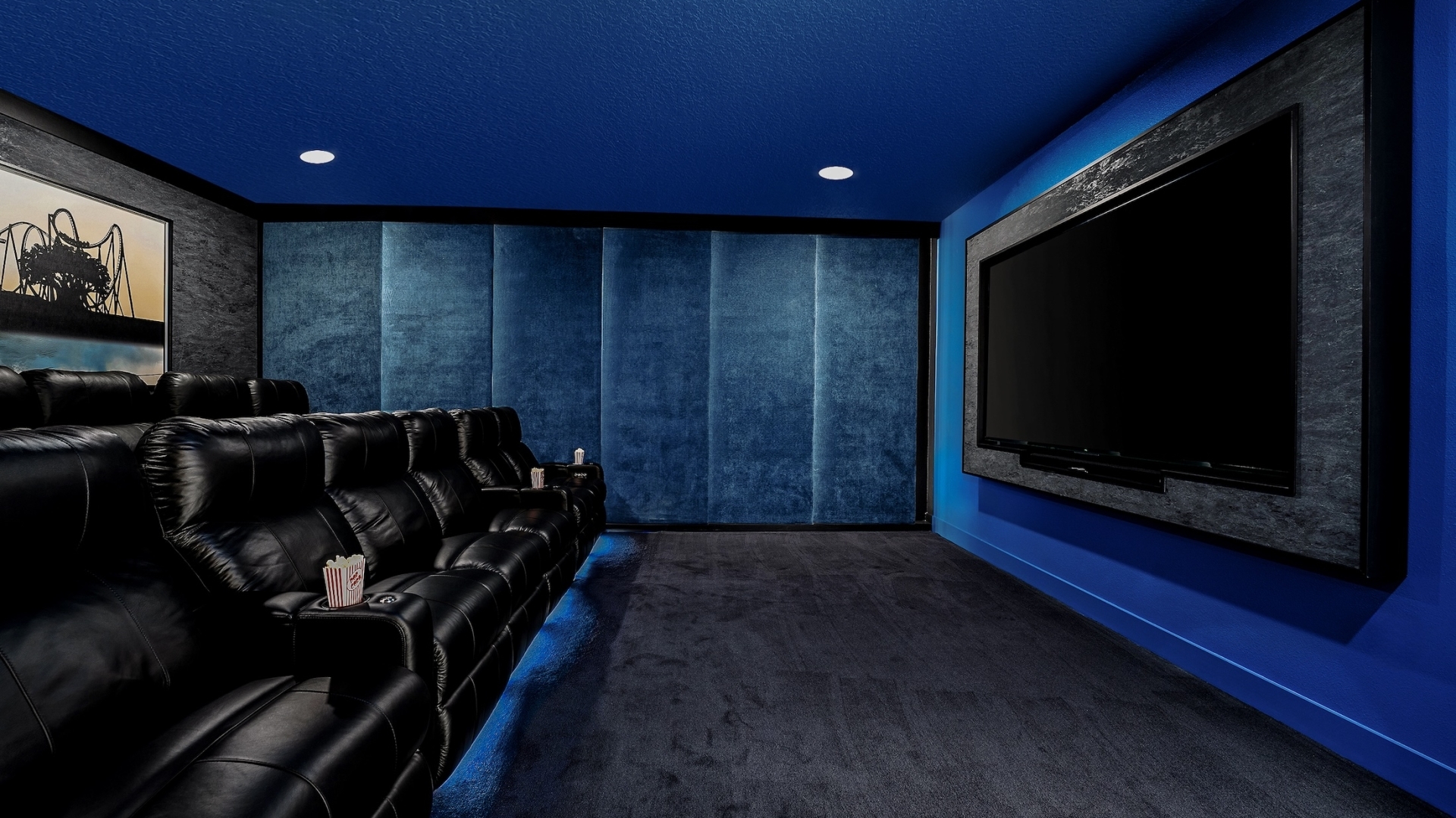 A private home theater room with tiered black leather recliners, a large wall-mounted screen framed in gray stone, and deep blue accent walls.