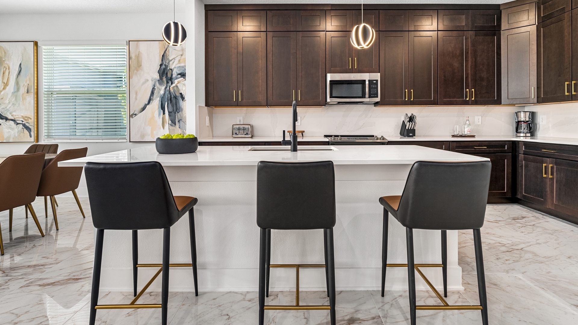 A spacious kitchen featuring minimalist light gray cabinets, a white quartz island with three black leather barstools, and polished tile flooring.