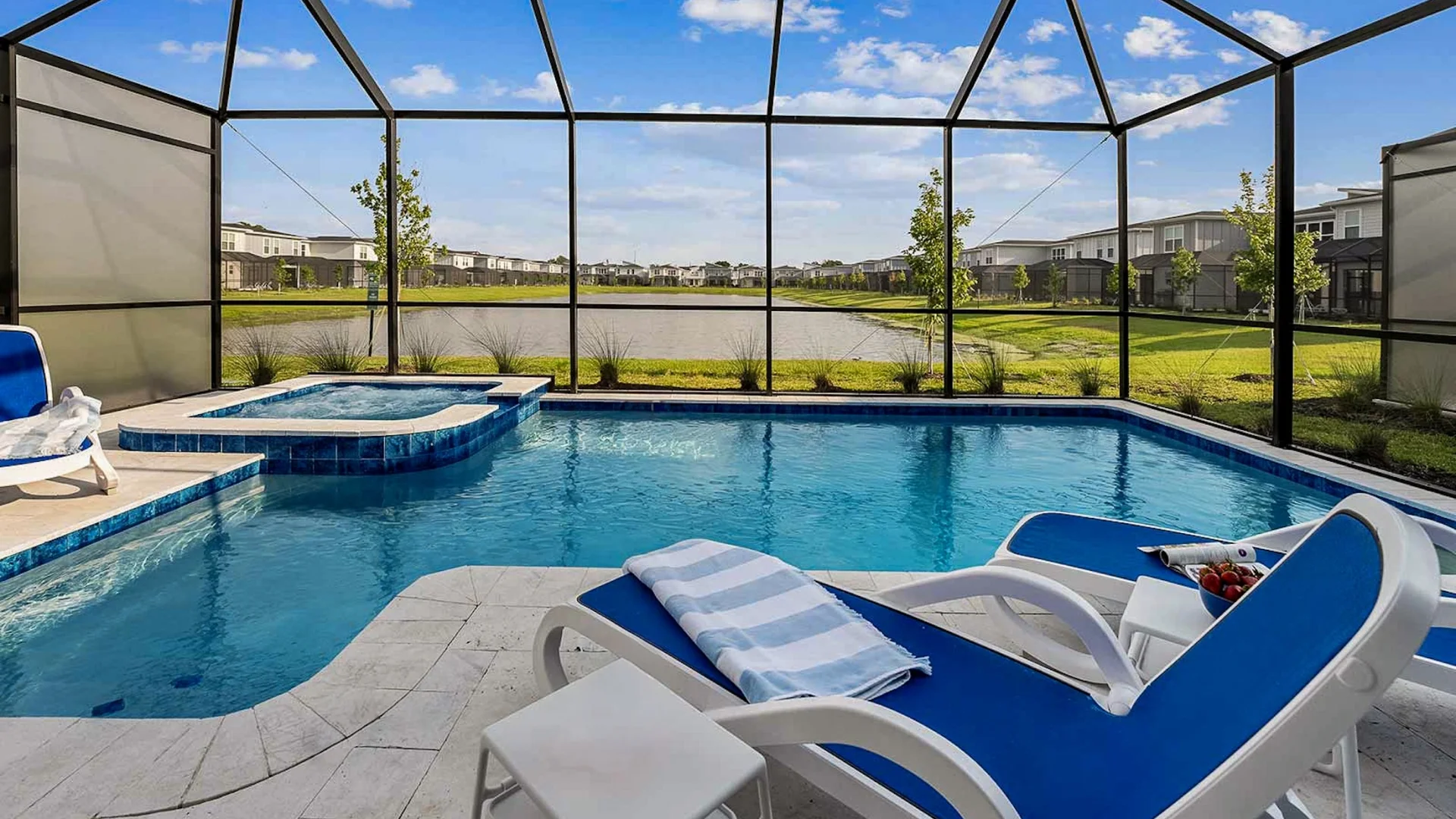A private screened-in patio featuring a swimming pool, integrated hot tub, and blue lounge chairs overlooking a scenic community lake.
