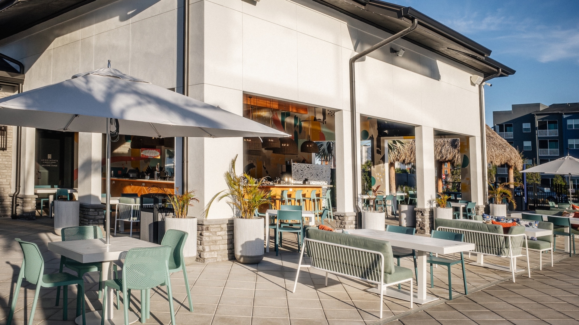 The outdoor patio of Aqua Bar & Grille featuring modern teal chairs, white tables with umbrellas, and cushioned lounge seating outside a white contemporary building.