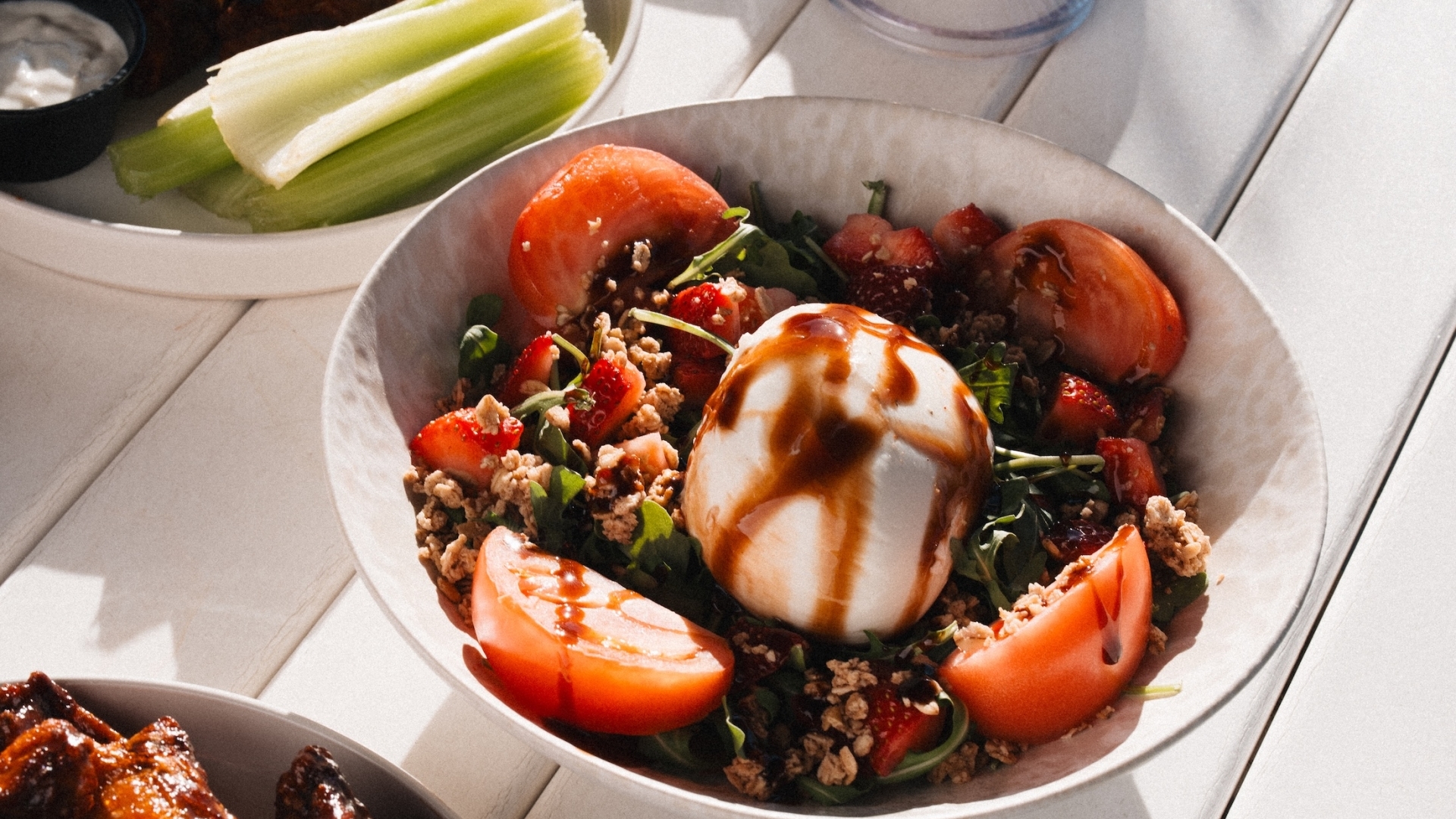 Close-up of a fresh salad with burrata cheese, tomatoes, and balsamic glaze, surrounded by other poolside dishes and drinks.