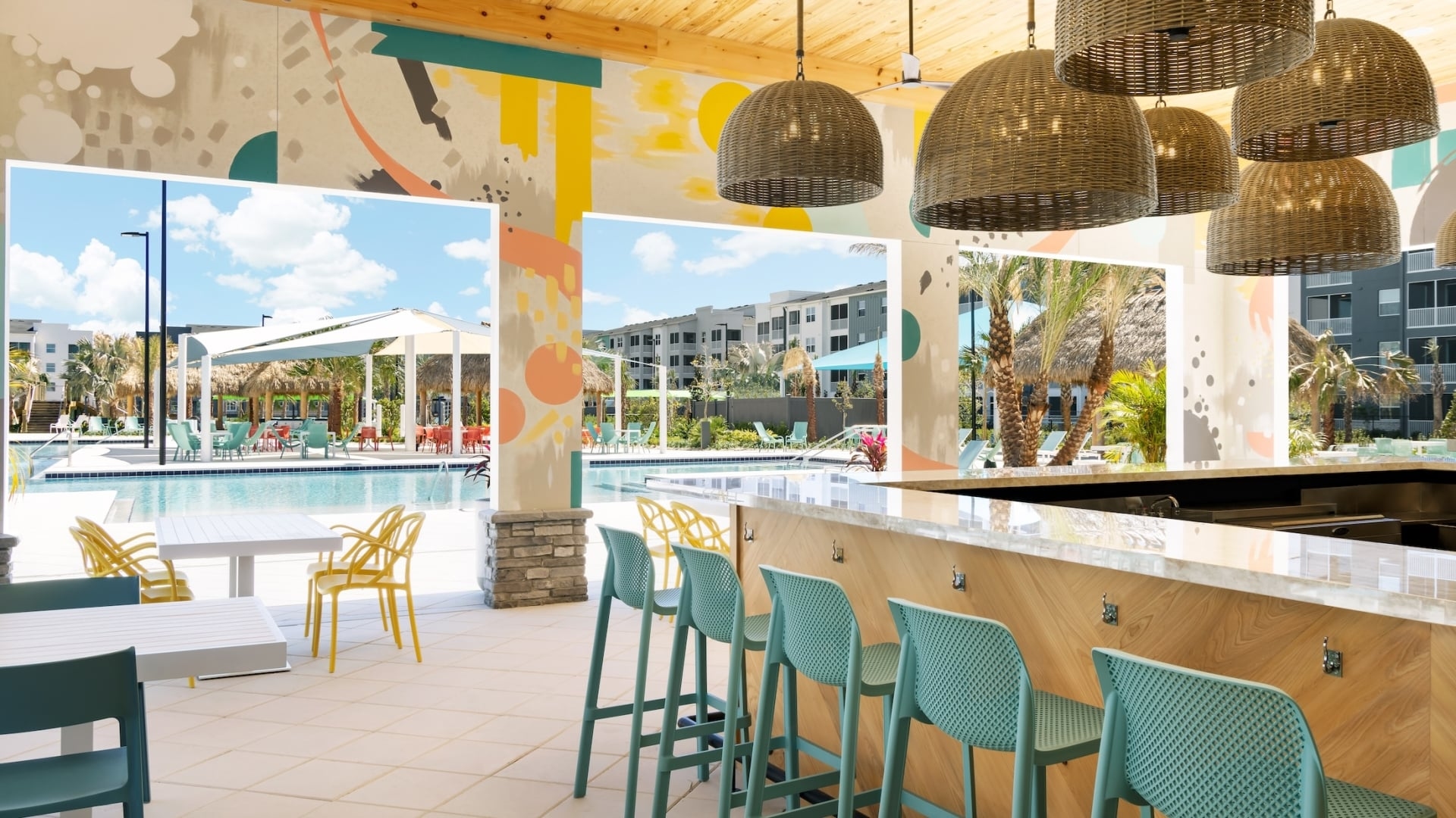 A vibrant poolside bar featuring teal barstools, woven pendant lights, and colorful mural-covered pillars overlooking a swimming pool.