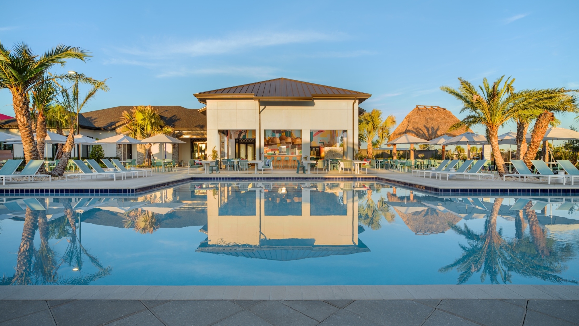 A calm resort swimming pool reflecting a modern clubhouse building and palm trees at sunset, with thatched-roof tiki huts and umbrellas in the background.