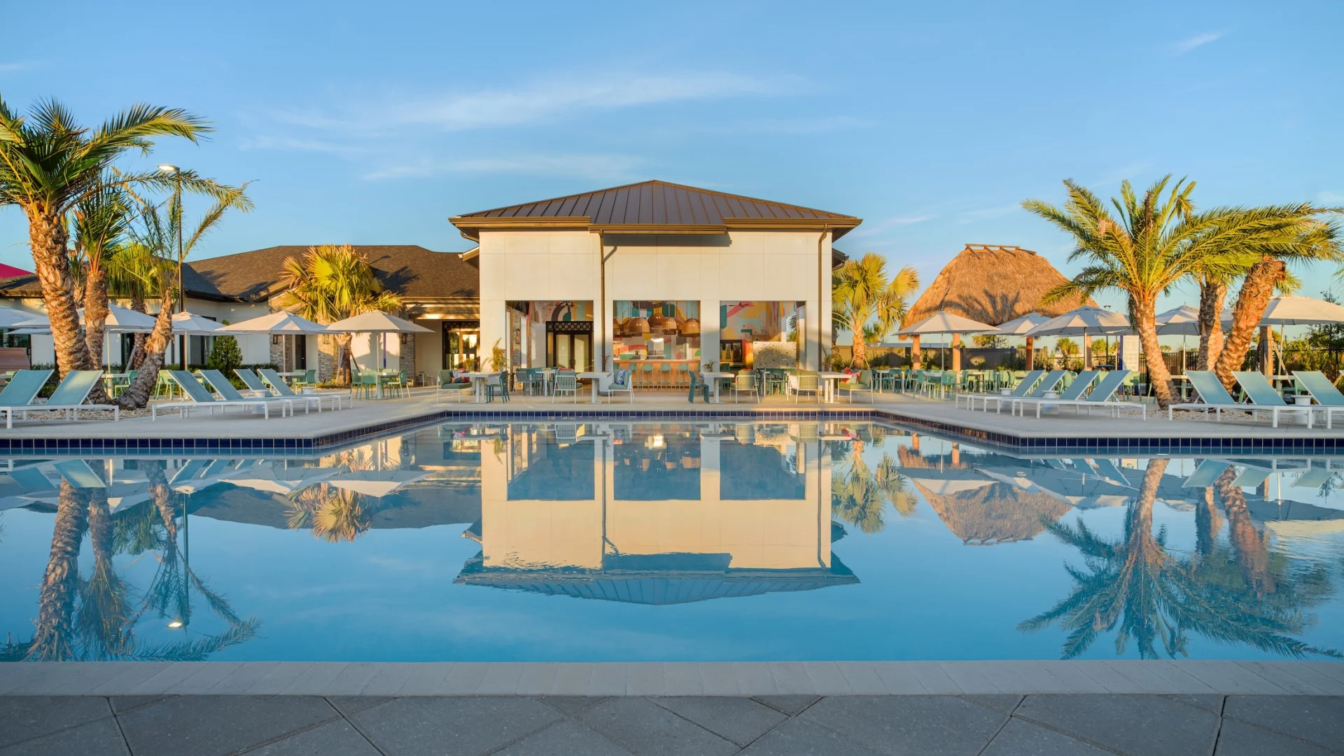 A calm resort swimming pool reflecting a modern clubhouse building and palm trees at sunset, with thatched-roof tiki huts and umbrellas in the background.