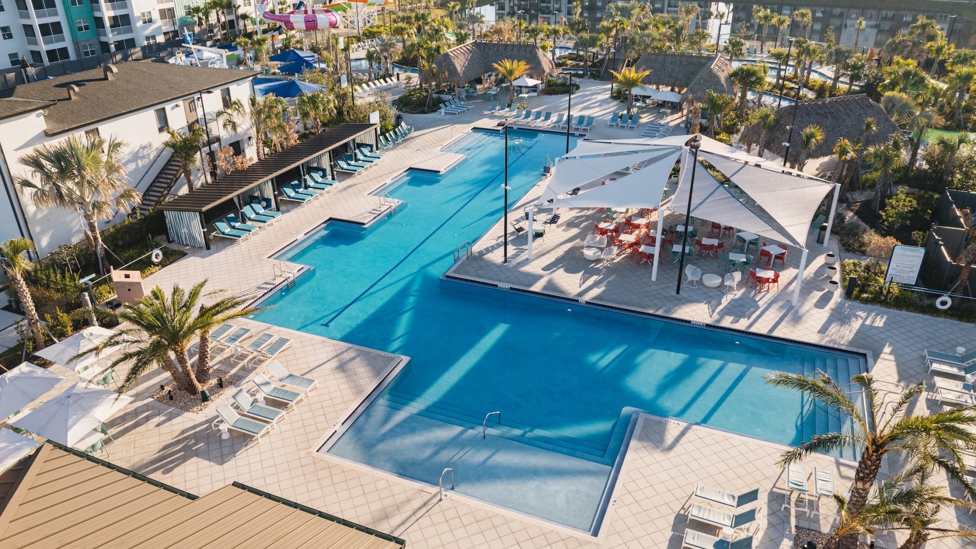 High-angle aerial view of a large resort swimming pool area with sun loungers, private cabanas, and a shaded outdoor dining terrace, with water slides in the distance.