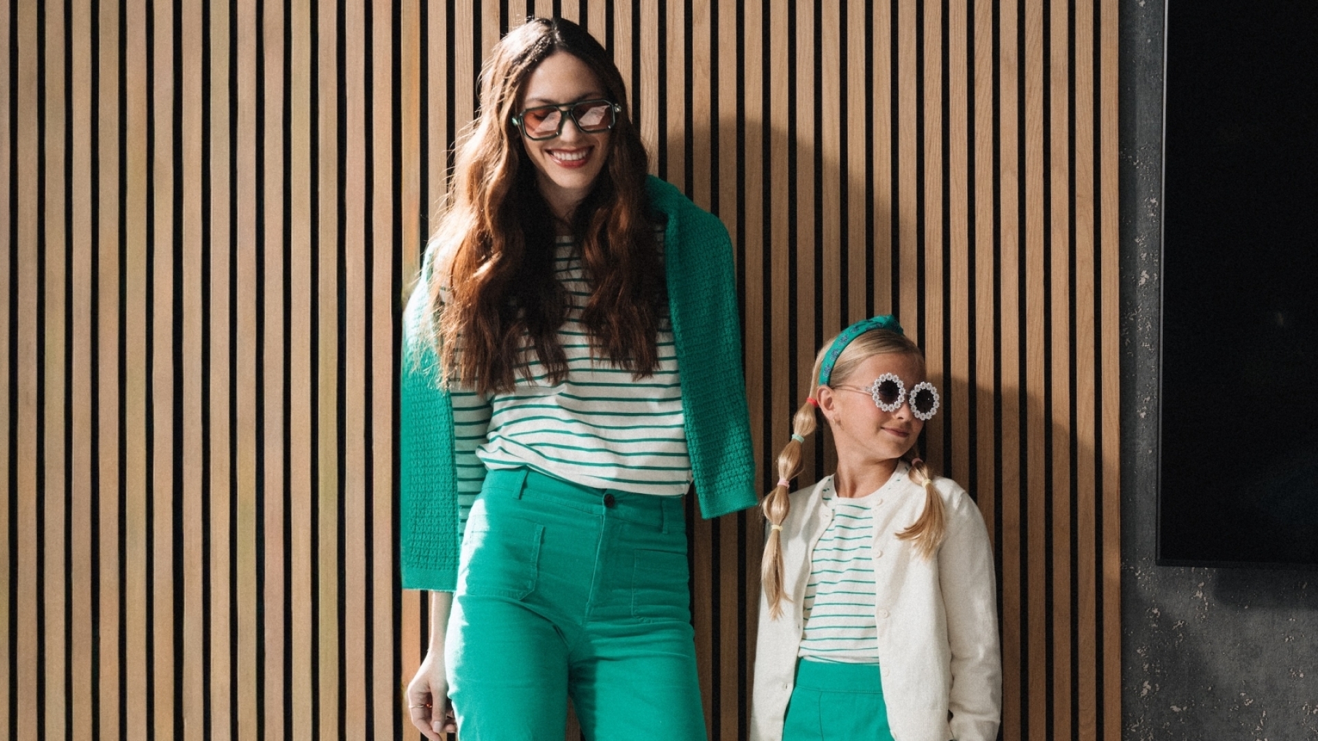 A mother and young daughter in coordinated green and white outfits posing together against a modern wooden slat wall inside a resort.