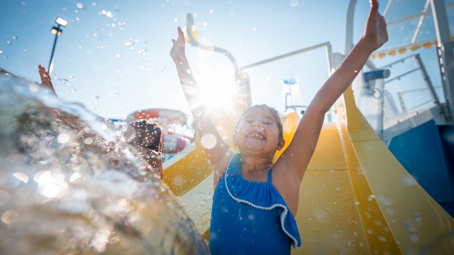A young girl in a blue swimsuit smiles with her arms raised in mid-air as she splashes into the water at the bottom of a bright yellow water slide on a sunny day.