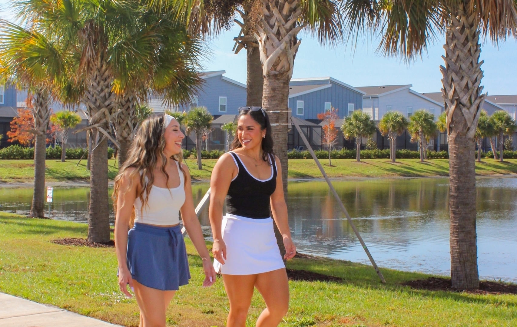 Two women in athletic wear walking and talking along a paved lakeside path lined with palm trees at a resort.