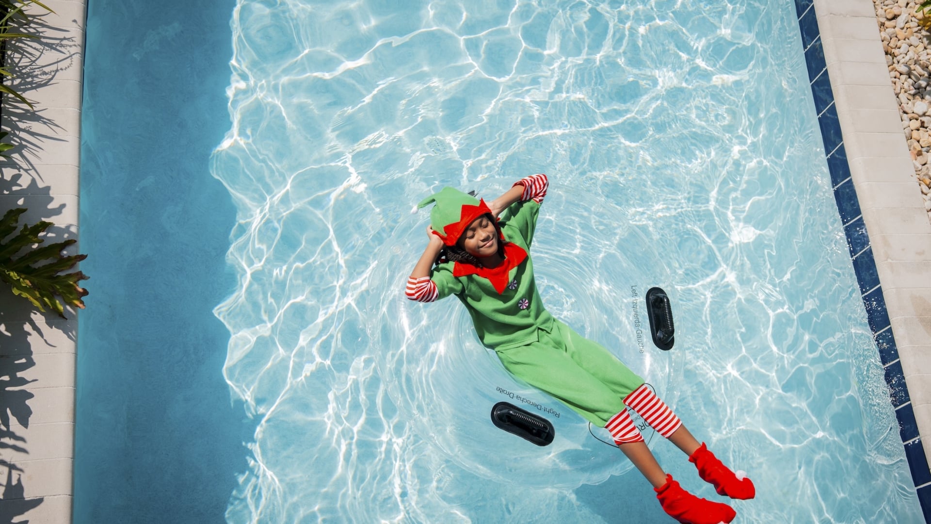 An overhead view of a child dressed in a green and red holiday elf costume floating on a clear inner tube in a bright blue swimming pool.