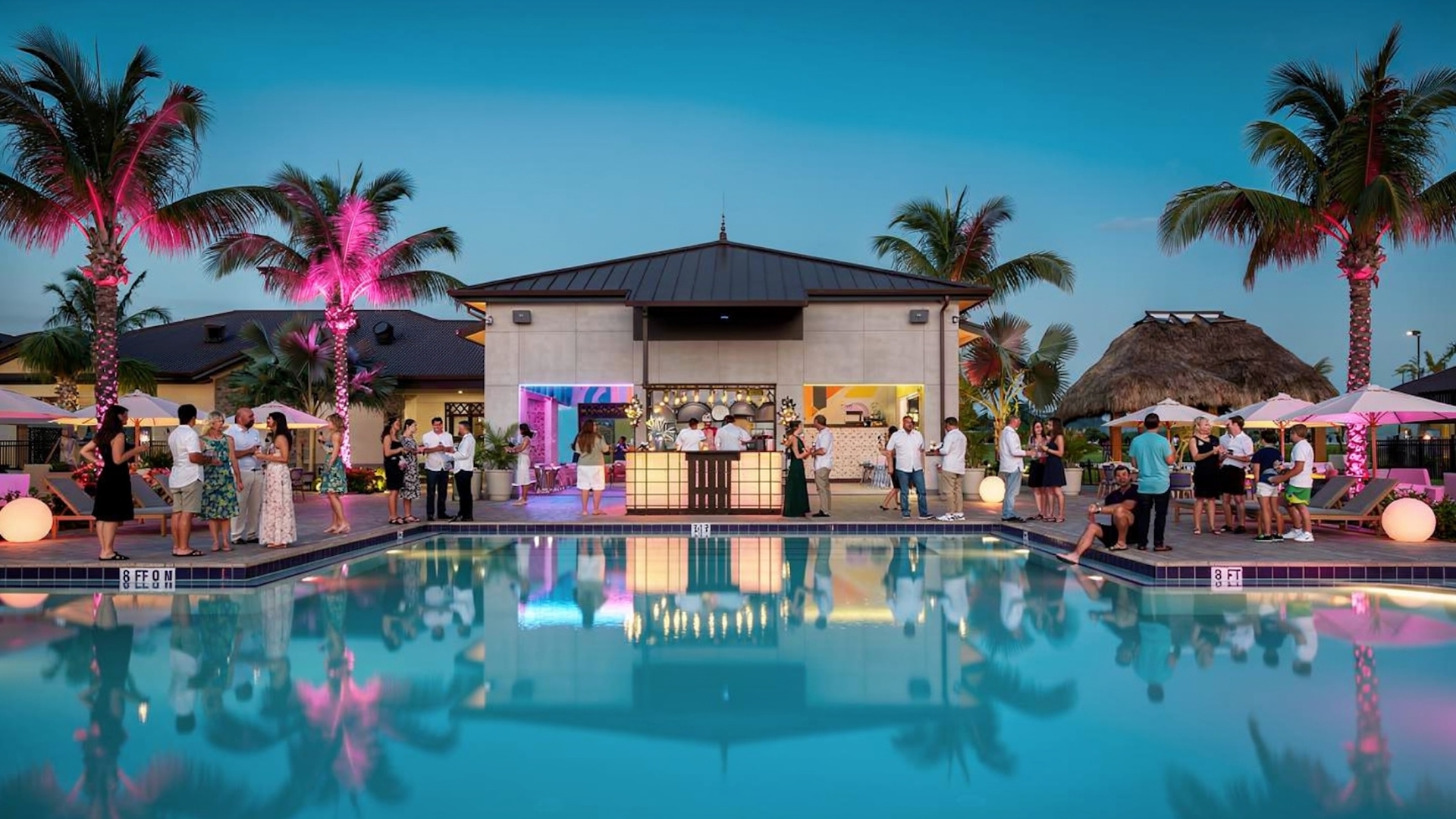 An evening poolside reception at a resort featuring guests in semi-formal attire socializing around a lit outdoor bar and pool with palm trees illuminated in pink.