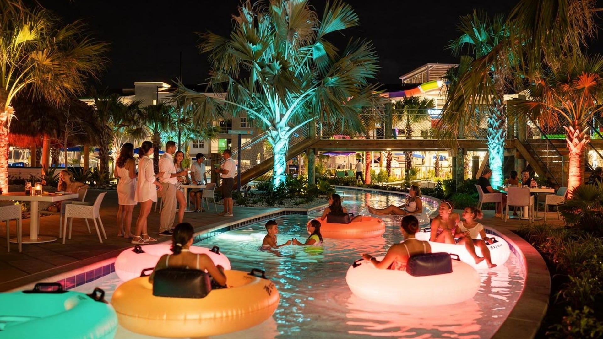 A vibrant nighttime scene at a resort lazy river featuring guests on glowing inner tubes, illuminated palm trees, and people dining at poolside tables.