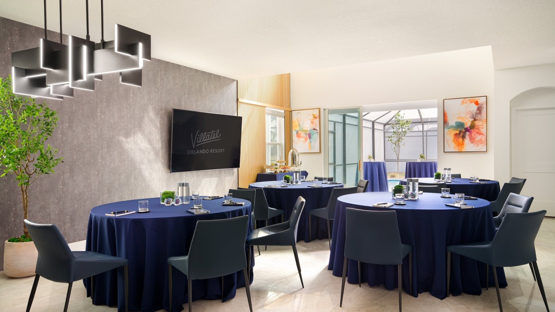 A modern living area repurposed as a meeting space with several round tables draped in navy blue linens and a wall-mounted display.