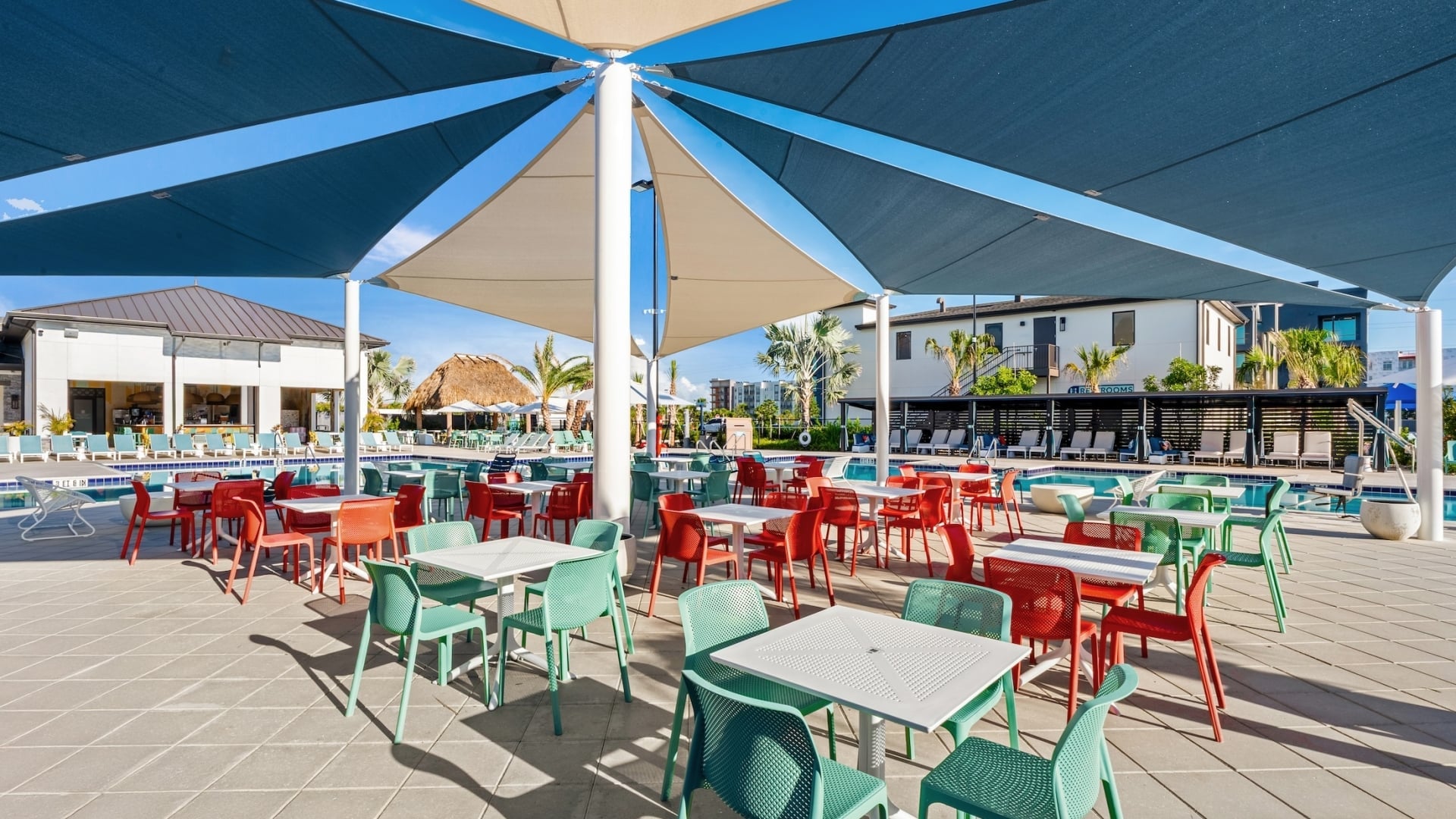 An outdoor event pavilion with red and teal seating arranged under large blue and tan sun sails next to a resort pool.