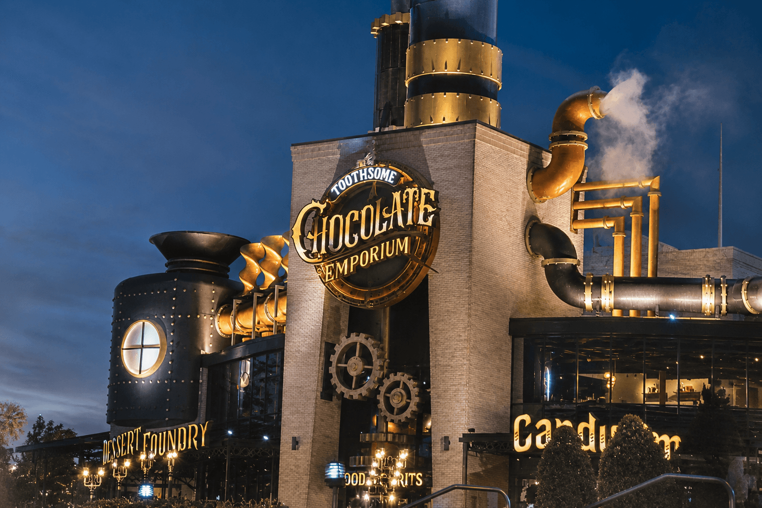 The illuminated steampunk-inspired facade of The Toothsome Chocolate Emporium at night, showcasing giant smoking smokestacks, mechanical gears, and golden piping.