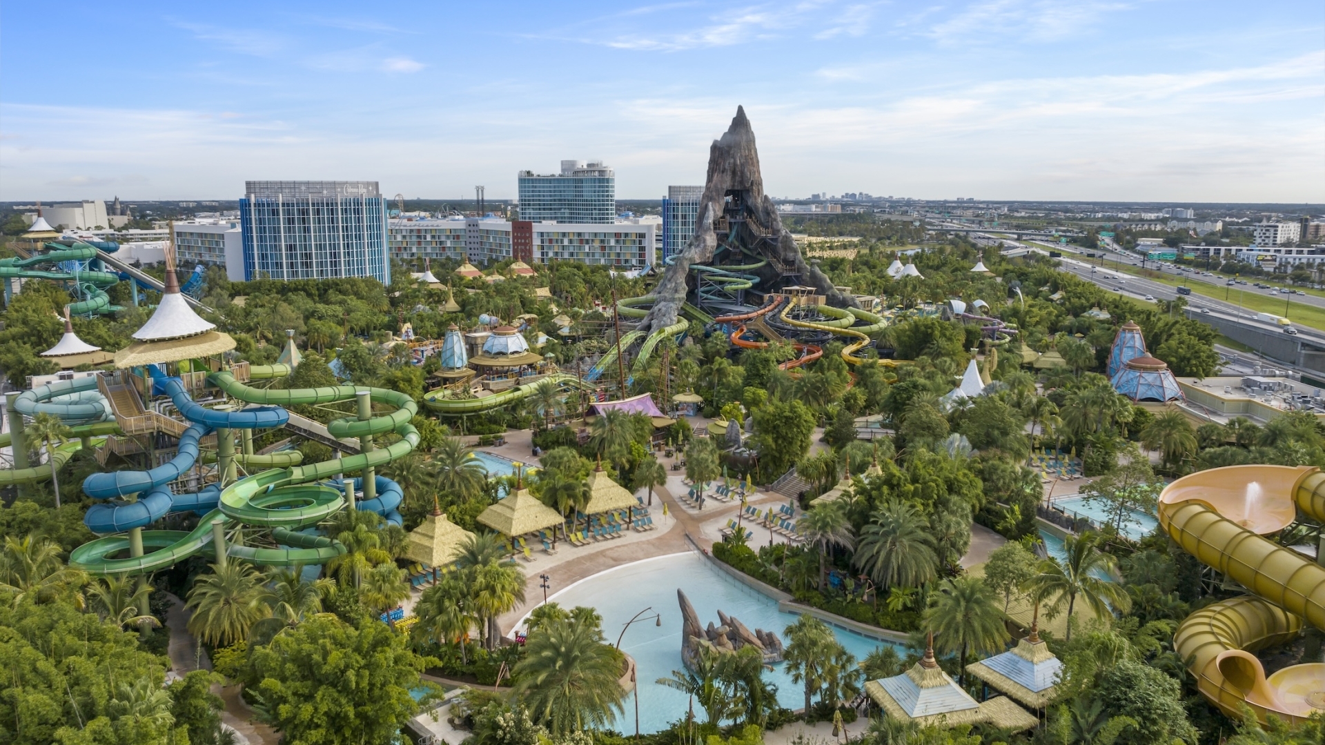 An elevated aerial view of Universal's Volcano Bay water theme park, highlighting the massive central Krakatau volcano surrounded by winding water slides, tropical trees, and sandy beach lagoons.