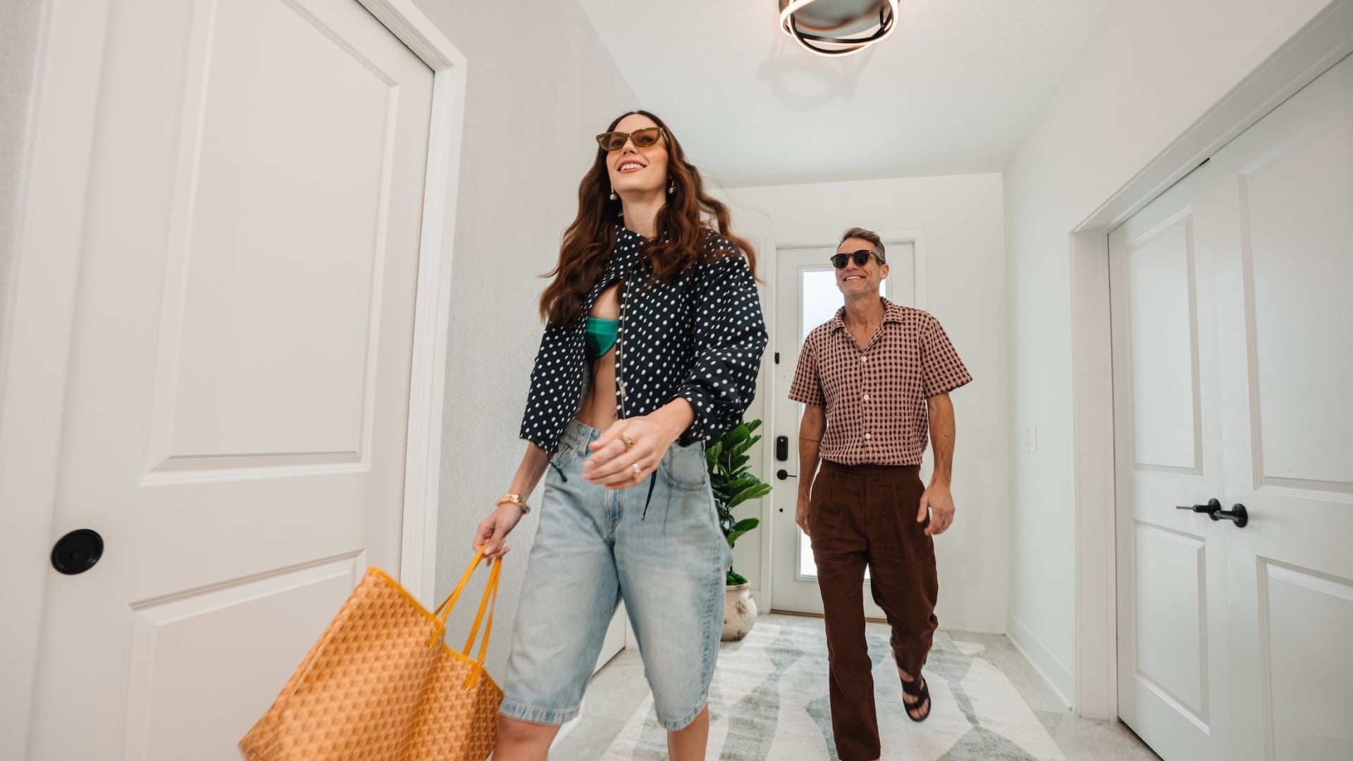 A stylish couple wearing sunglasses and summer attire walks through a bright, modern hallway of a vacation rental.