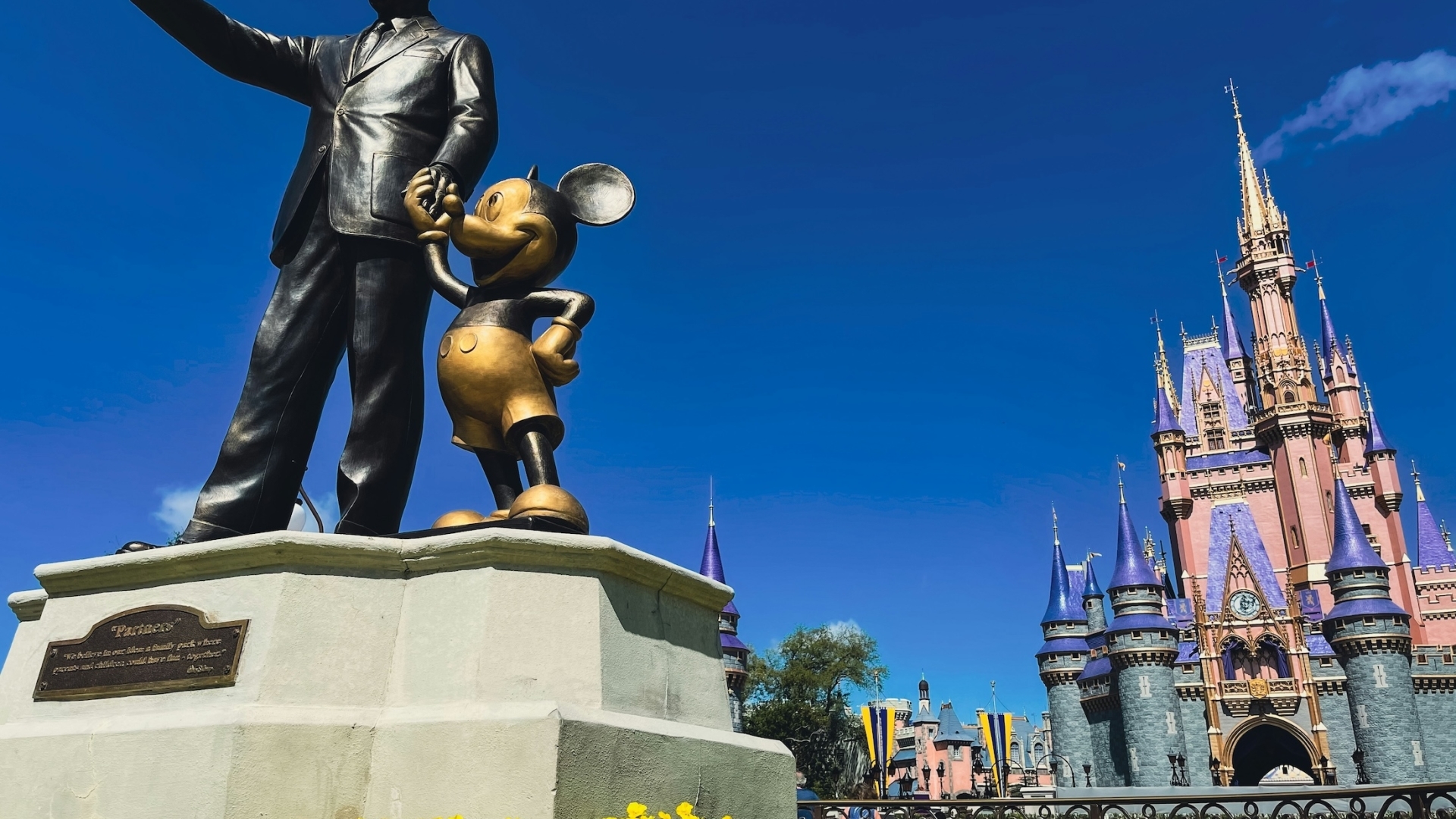 The bronze "Partners" statue of Walt Disney holding Mickey Mouse's hand stands in the foreground, surrounded by yellow flowers, with Cinderella Castle rising under a clear blue sky in the background.