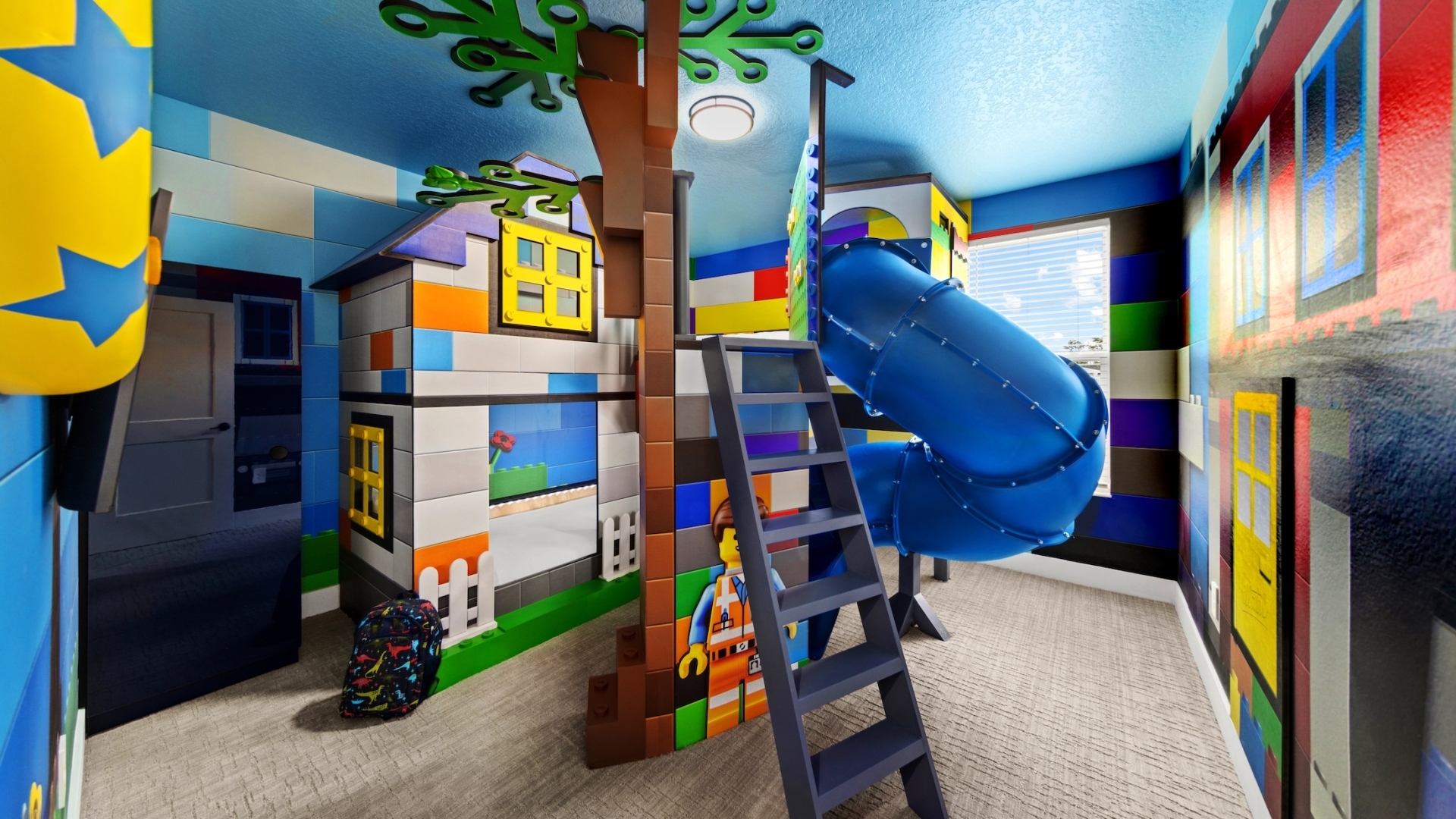 A colorful toy-brick-themed children's bedroom featuring a house-shaped bunk bed, a winding blue tunnel slide, and wall murals made to look like oversized building blocks.
