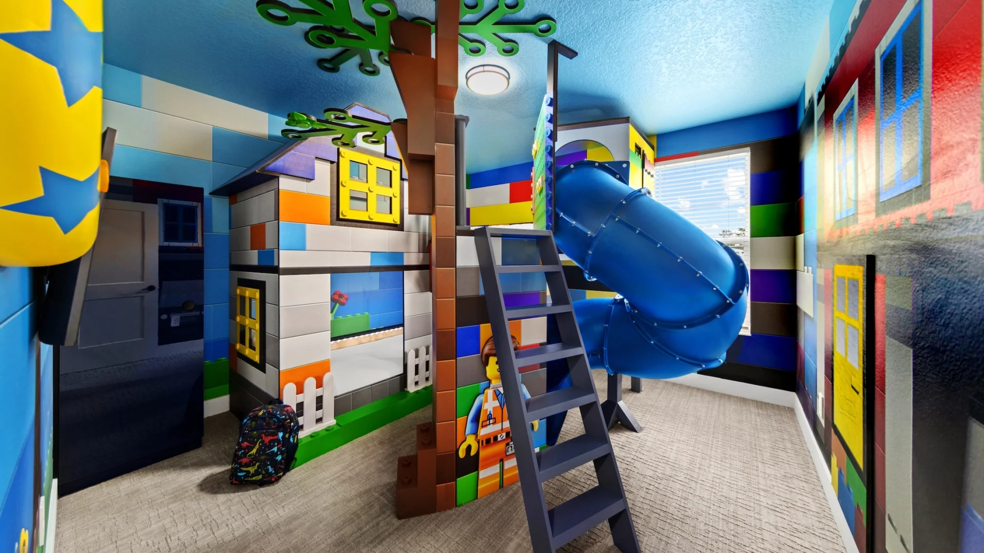 A colorful toy-brick-themed children's bedroom featuring a house-shaped bunk bed, a winding blue tunnel slide, and wall murals made to look like oversized building blocks.