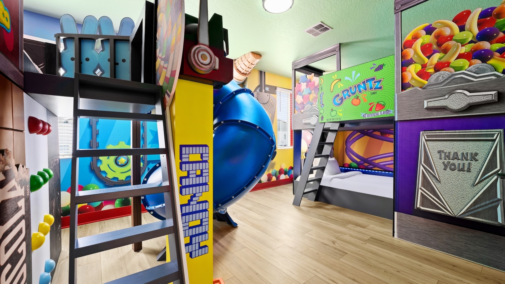 An elaborately themed children's bedroom featuring candy-inspired decor, custom bunk beds with a blue tunnel slide, and wall murals of gumball machines and sweets.