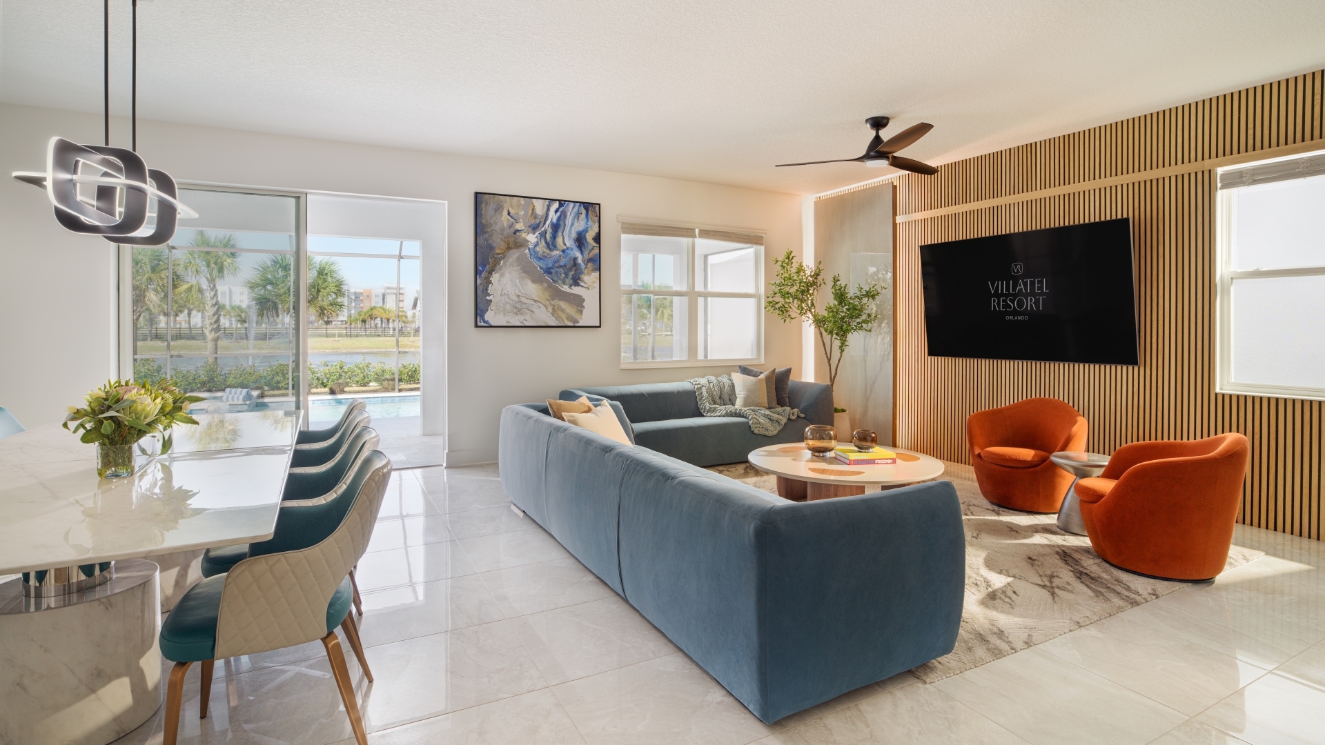 An open-concept villa interior showcasing a dining area with a marble table and brown chairs, a sleek blue sectional sofa set, and glass doors opening to a private pool courtyard.